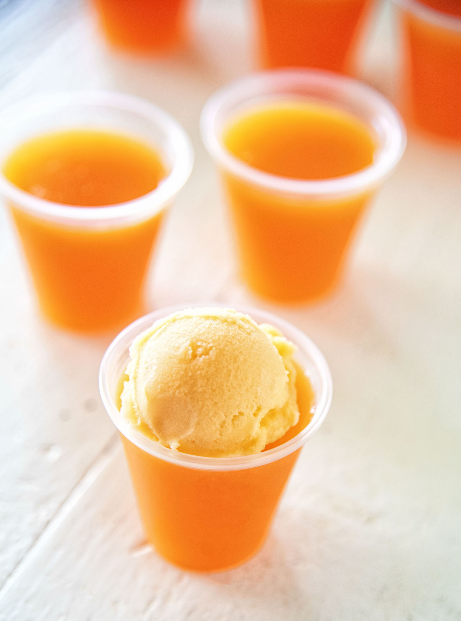 Several Orange Sherbet Jello Shots with the shot in focus topped with a mini scoop of orange sherbet. 
