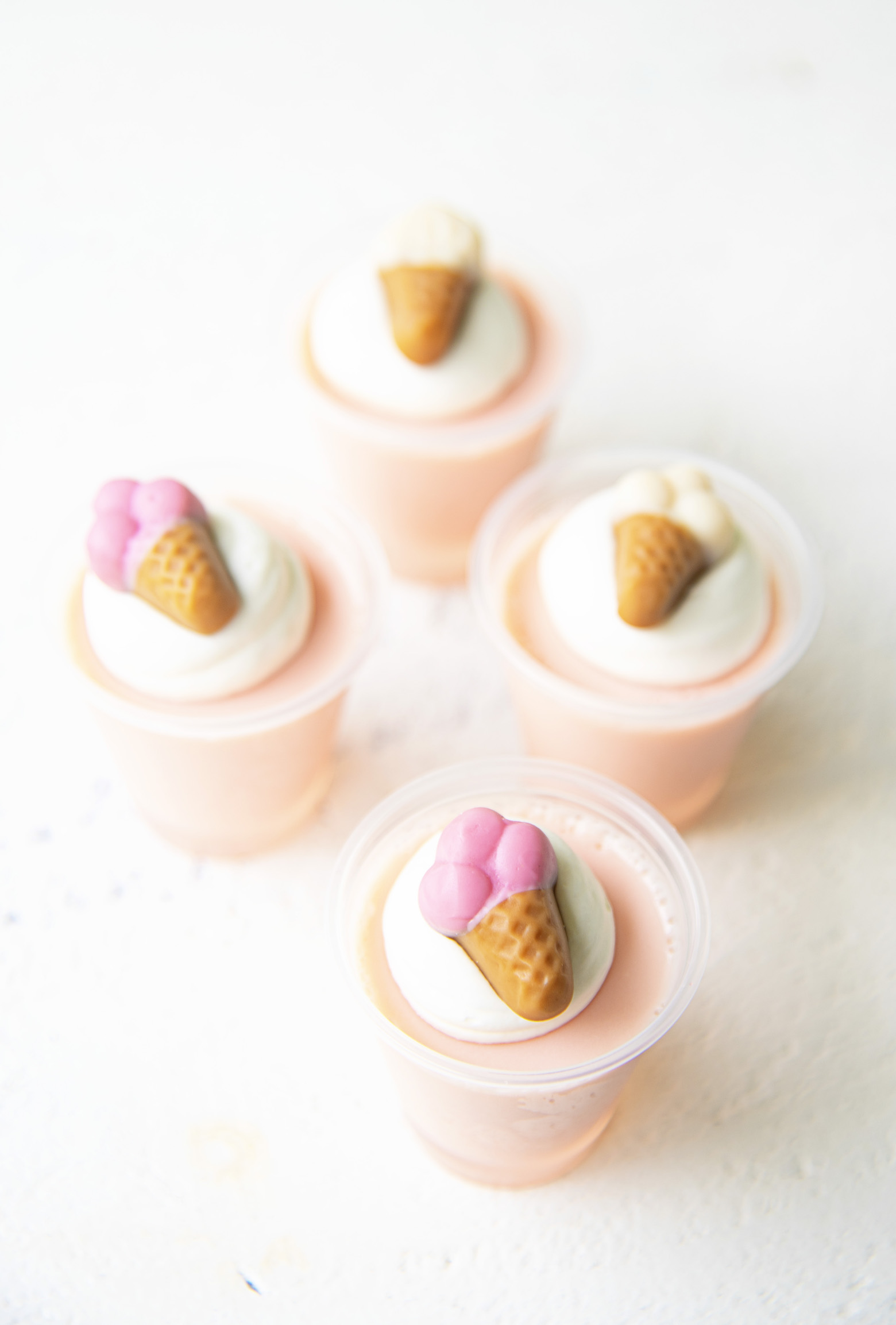 Four Strawberry Ice Cream Jello Shots with the focus on the first one. All shots are topped with whipped cream and mini gummy ice cream cones.