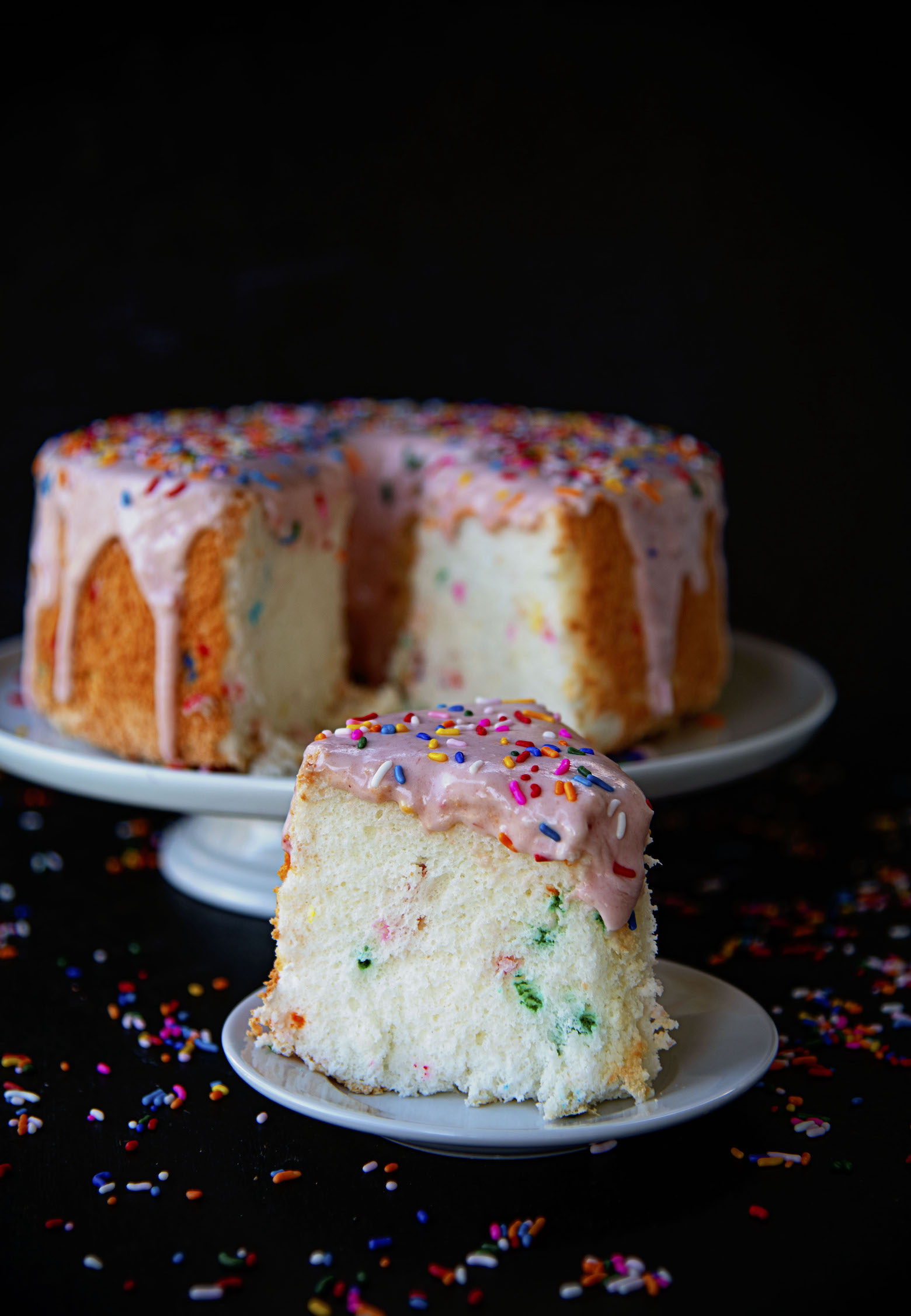 Slice of cake on a plate with the remaining Cake Batter Angel Food Cake in the background. 