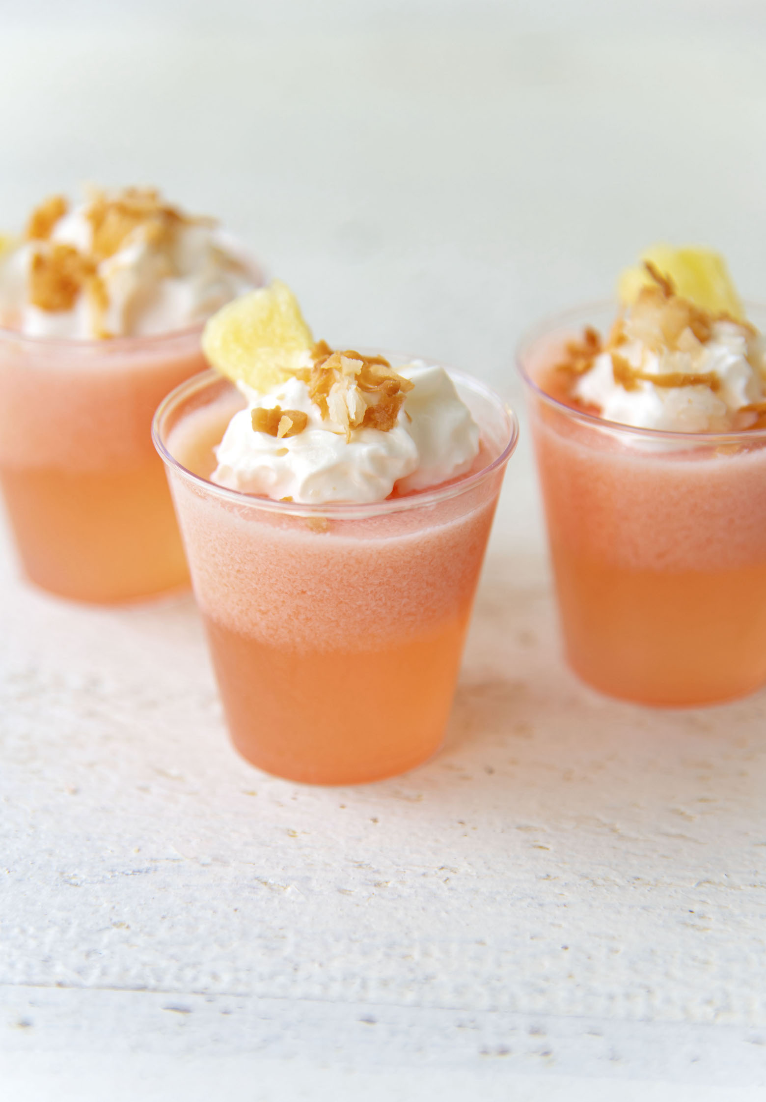 Three Strawberry Pina Colada Jello Shots with whipped cream, toasted coconut, and fresh pineapple on top.