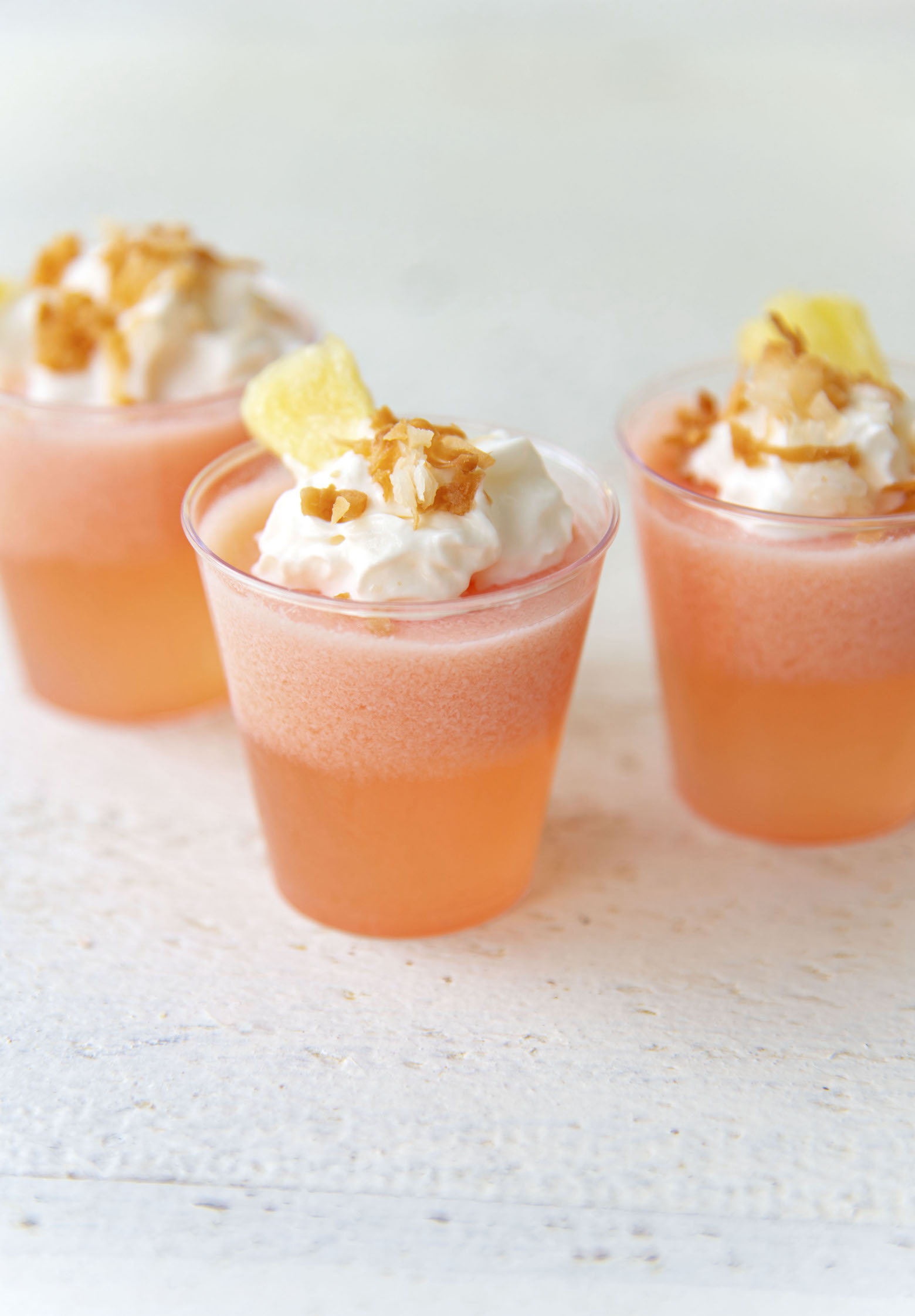 Three Strawberry Pina Colada Jello Shots with whipped cream, toasted coconut, and fresh pineapple on top. 