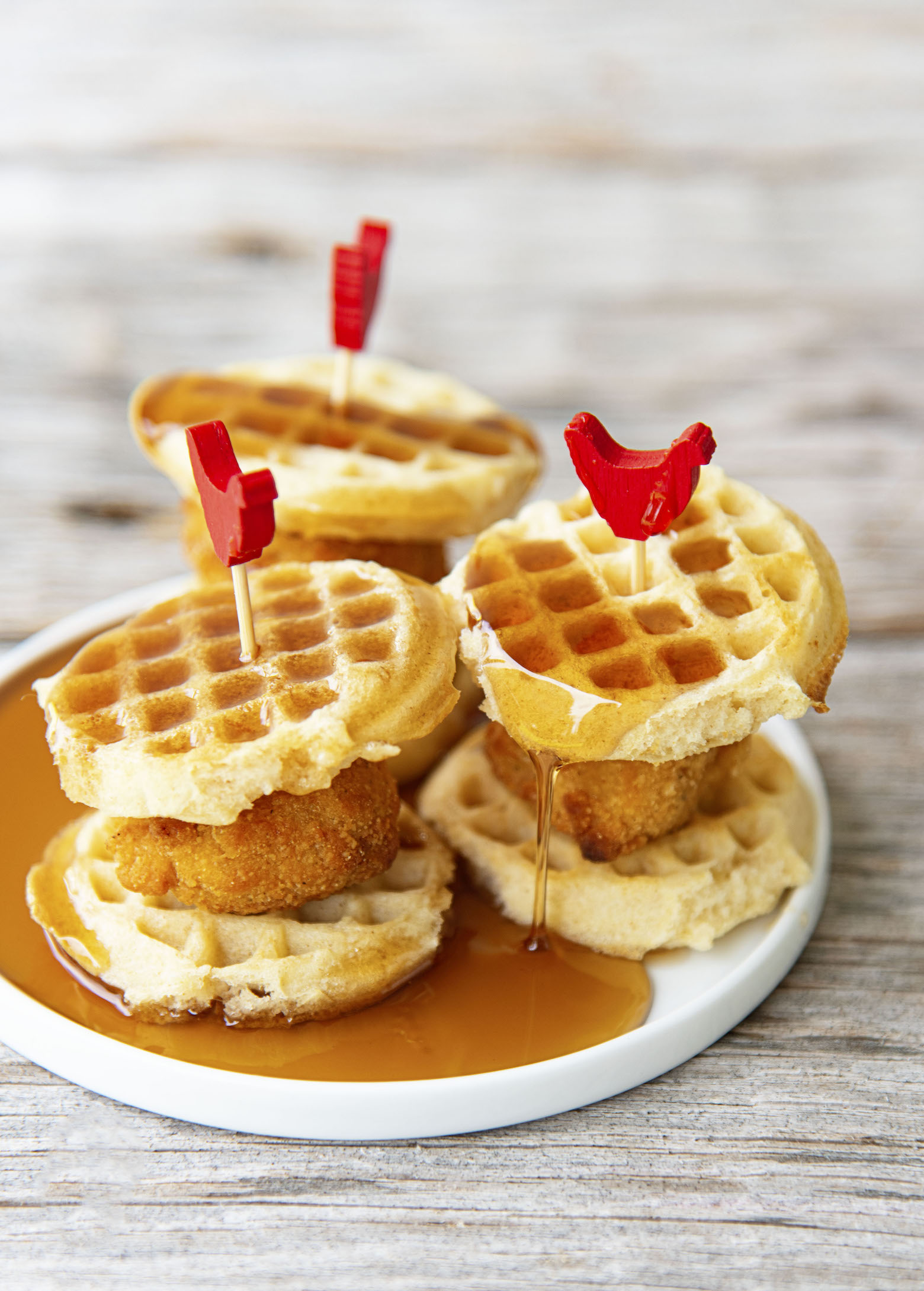 Three quarter shot of three Mini Chick’n and Waffles with syrup on them on a small plate. 