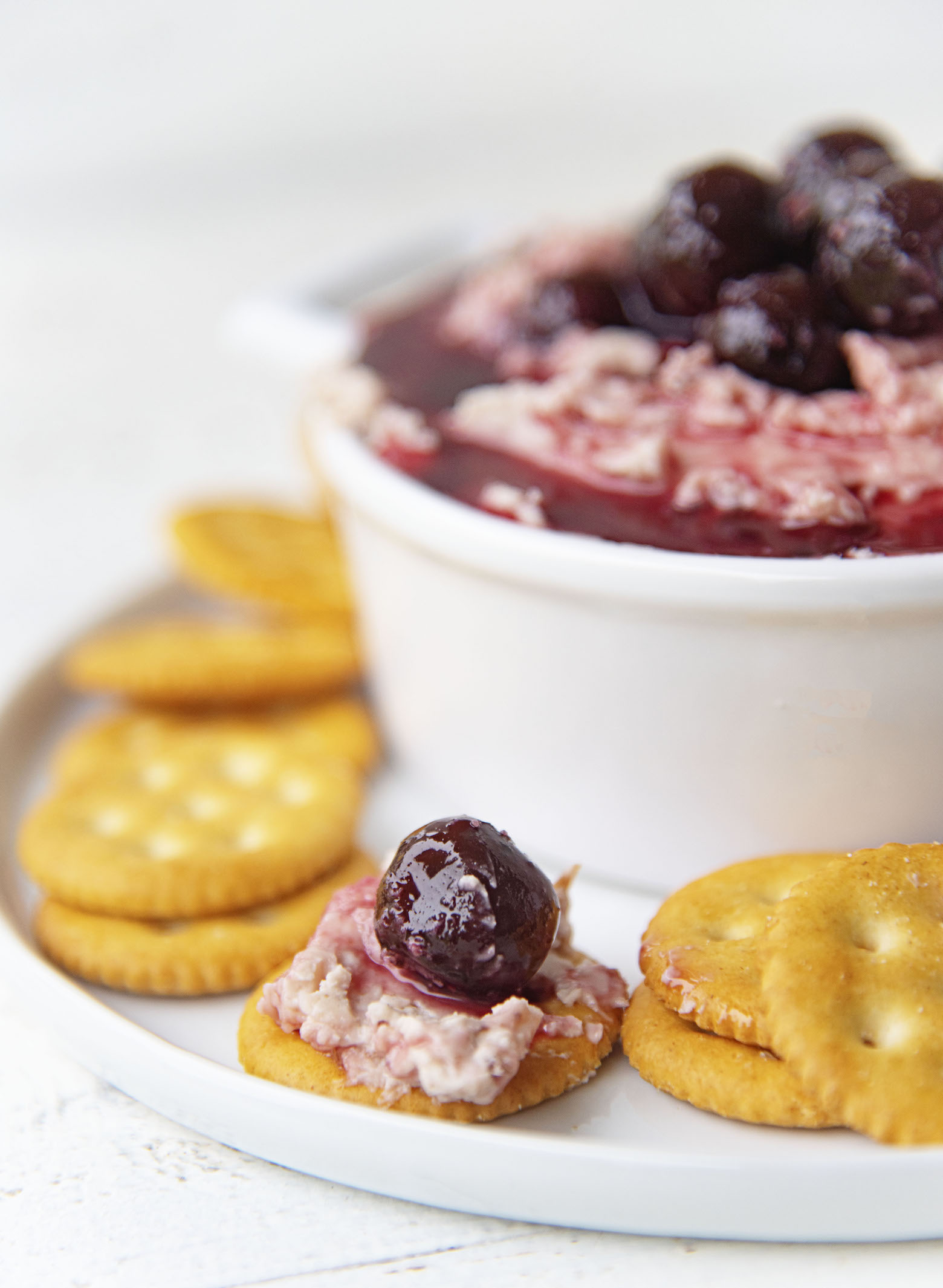 Side view of cracker with cheese spread and cherry on the side of the Cherry Cheddar Cheese Spread filled crock. 