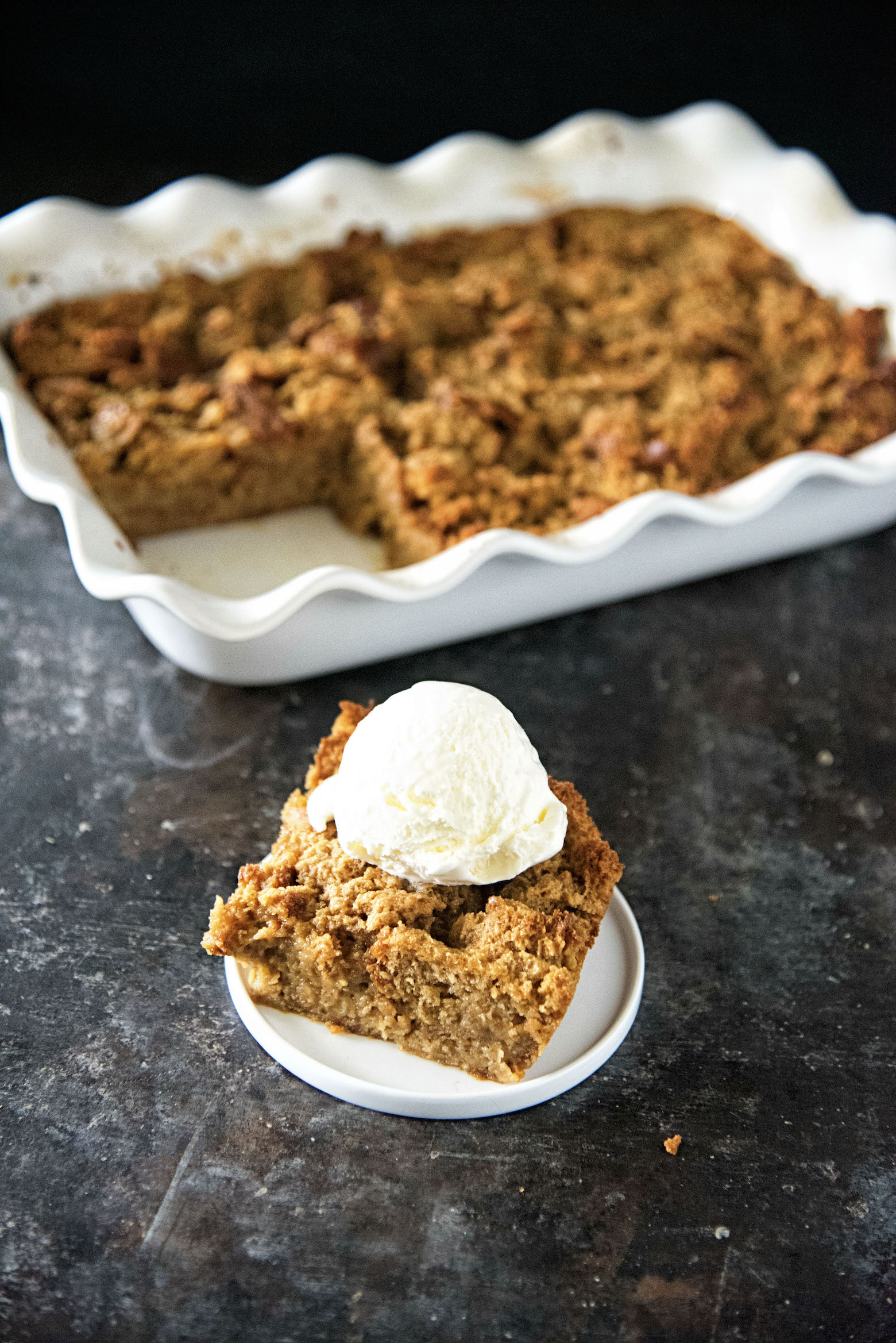 Slice of gingerbread bread pudding on a plate with ice cream on top with bread pudding pan in background. 