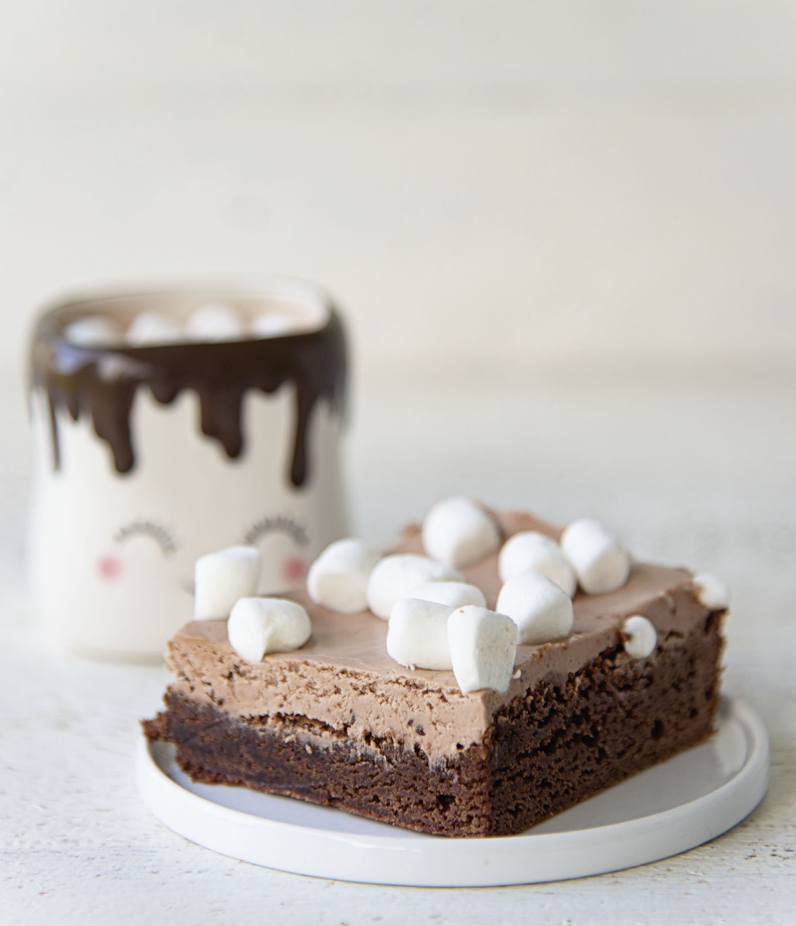 Side view of hot chocolate nougat brownie on a plate with cup of hot chocolate in the back. 