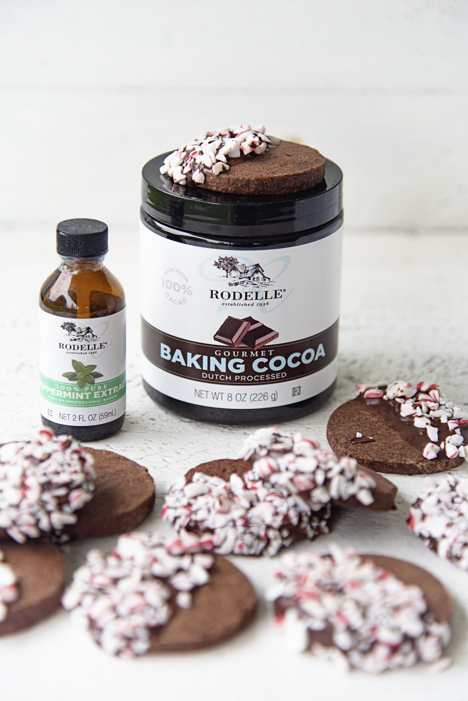 Scattering of peppermint mocha shortbread cookies with Rodelle Baking Cocoa and Peppermint Extract in the background.