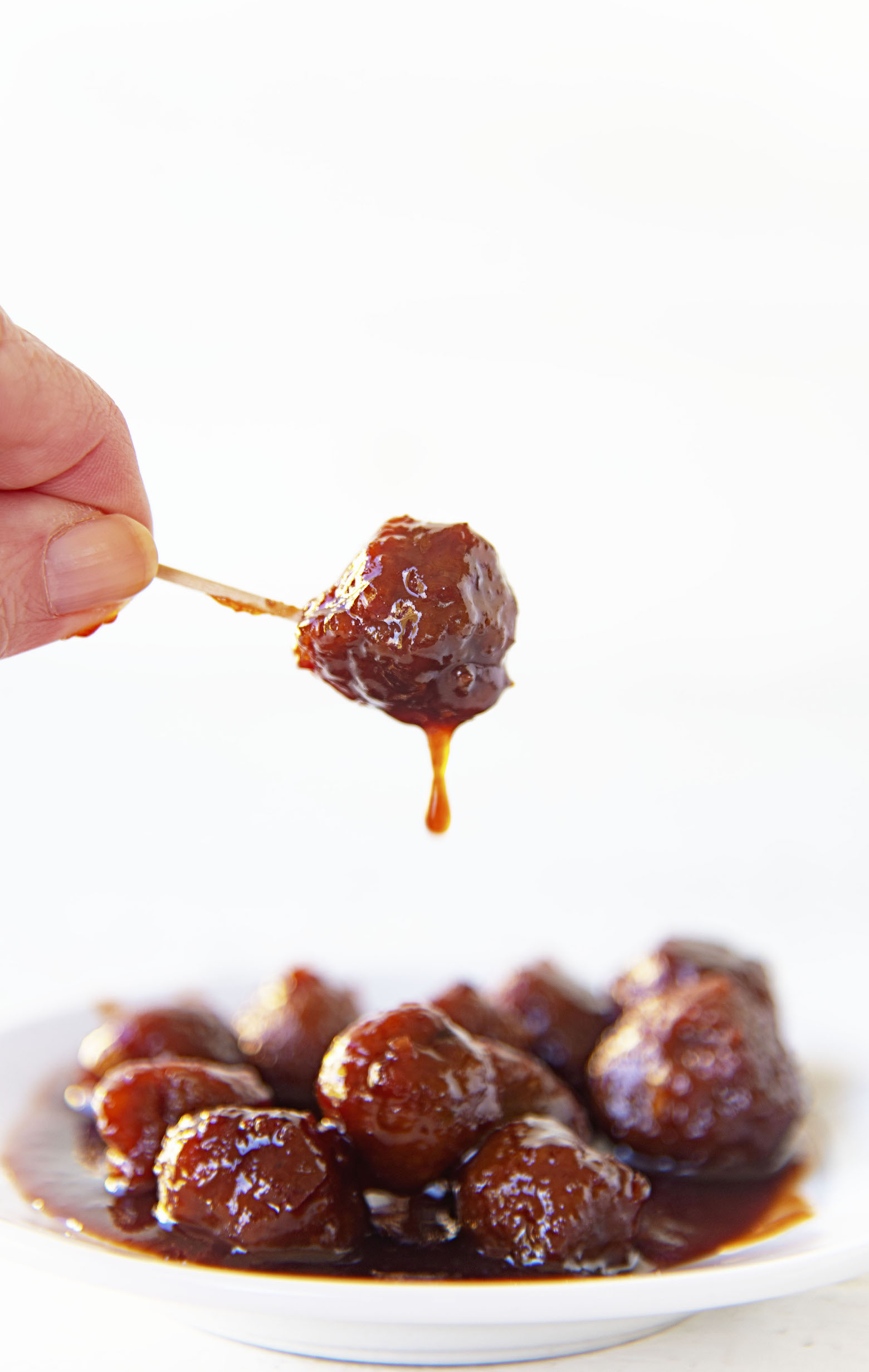Single meatball on a toothpick being held up from a plate of meatballs with sauce dripping off the single meatball. 