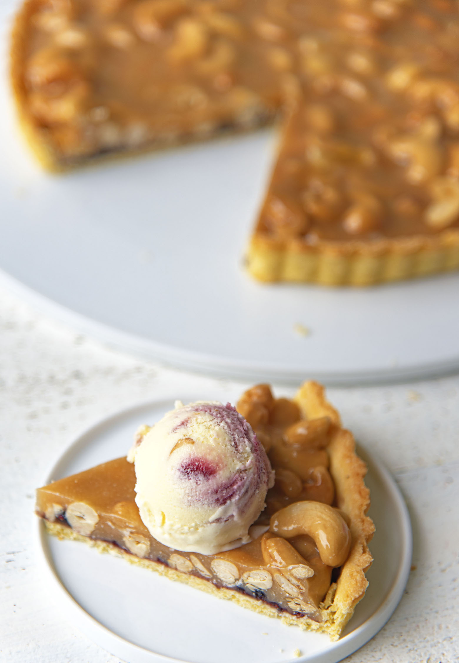 Slice of Baseball Nut Caramel Tart on a small plate with ice cream scoop on top with remaining tart in the background. 