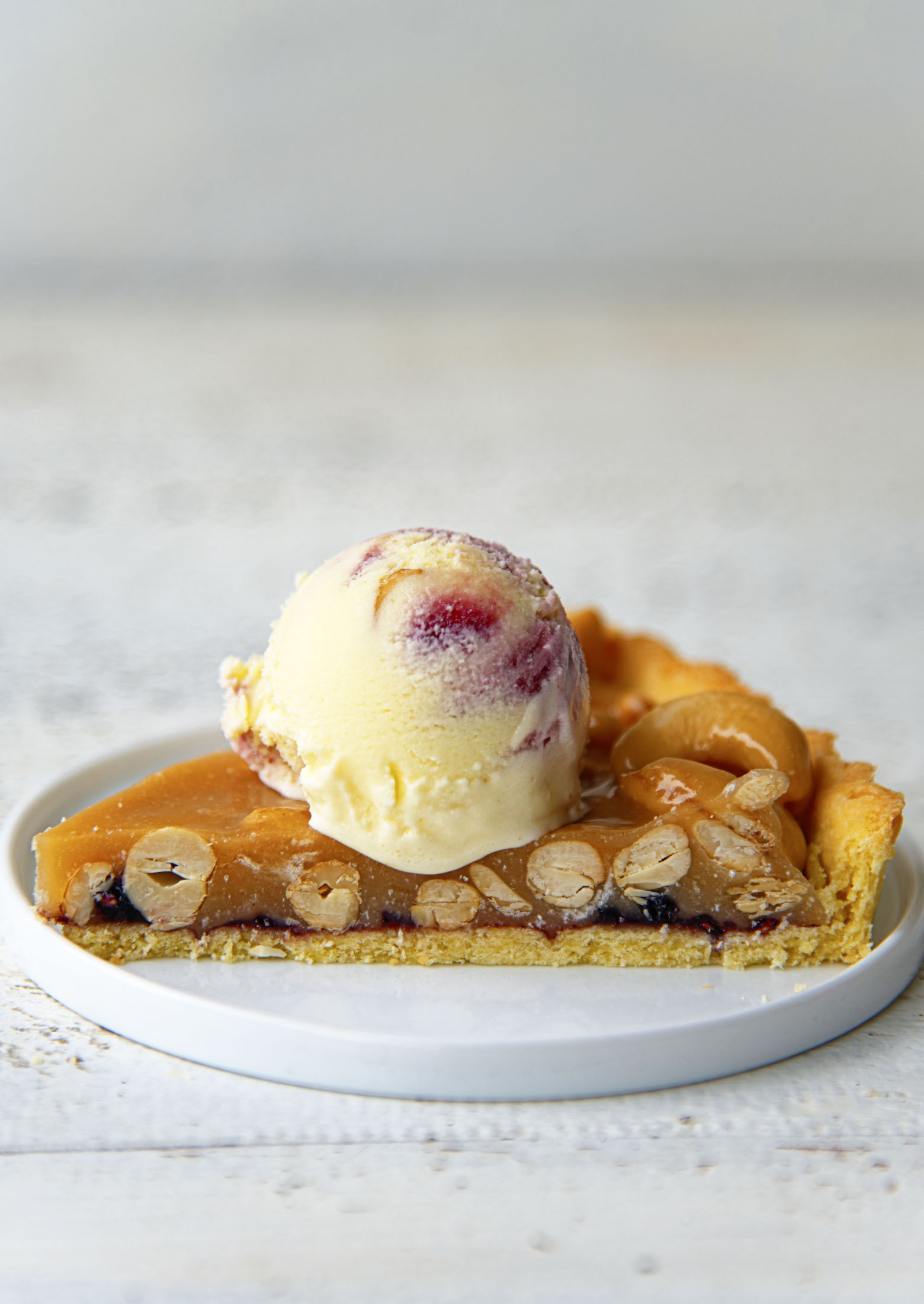 Side view of a piece of tart with a cross section exposing the cashews, jam layer, and bottom of tart. All topped with a scoop of ice cream. 