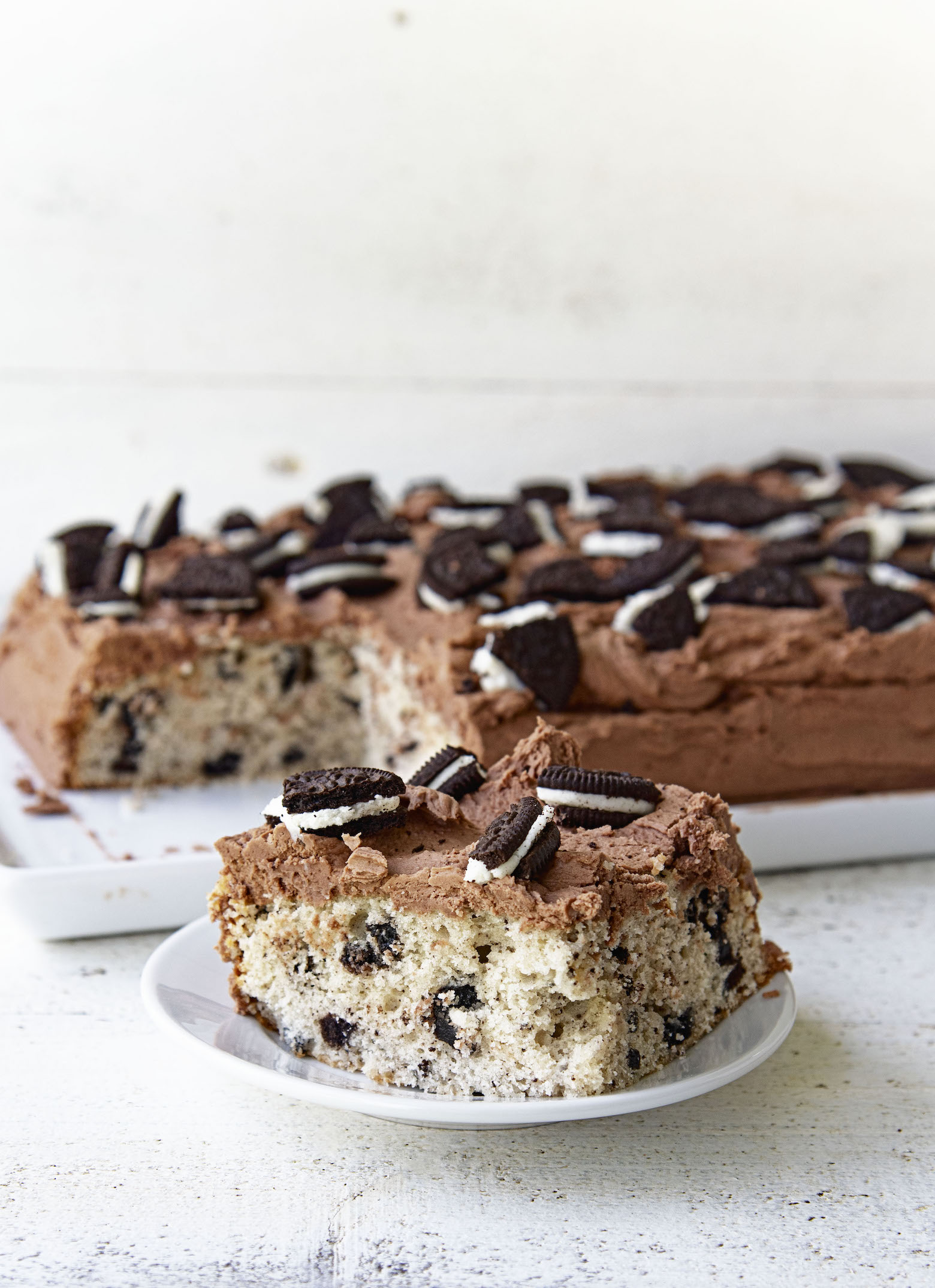 Piece of Cookies and Cream Cake on a plate with remaining cake in the background.