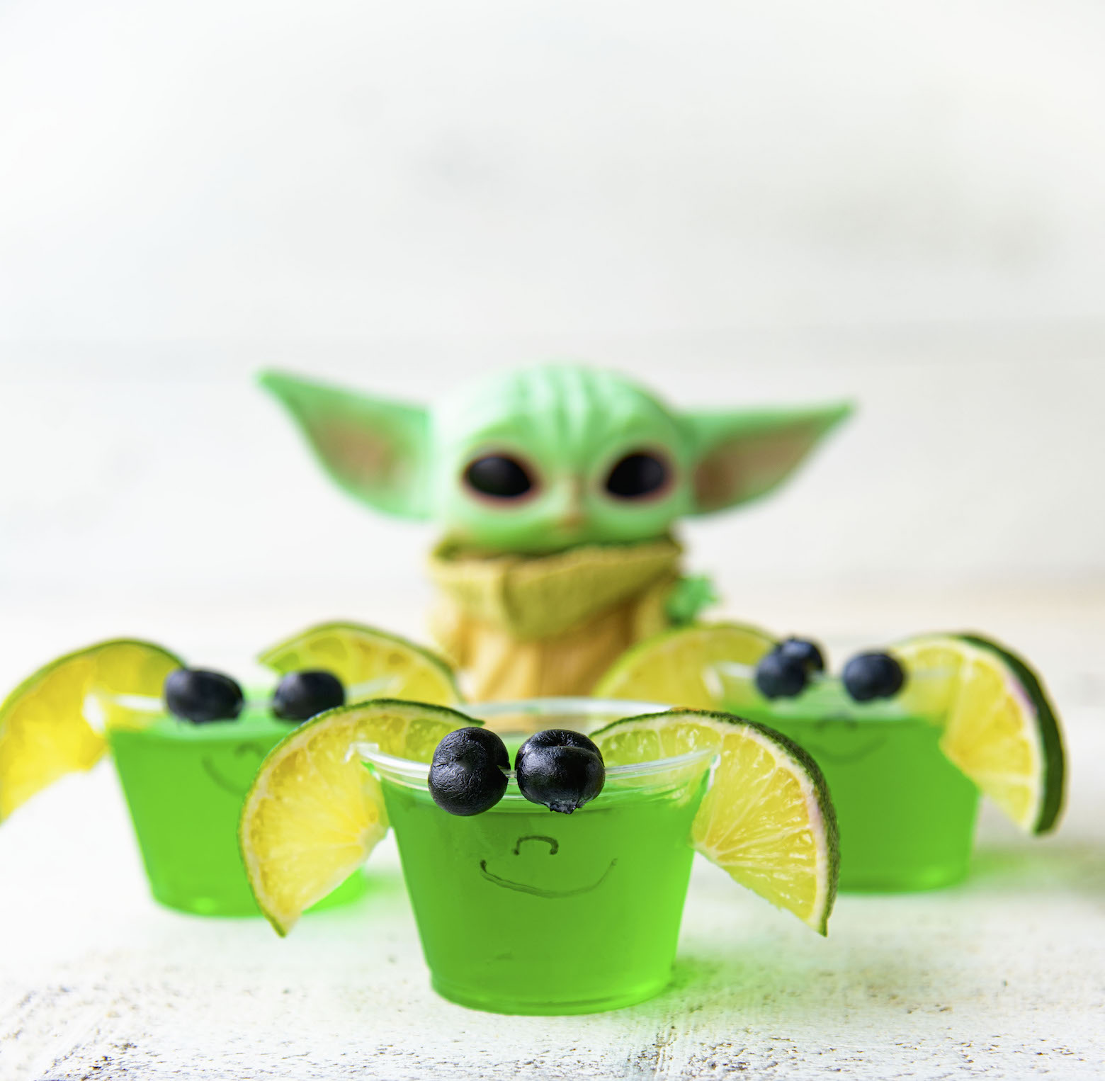 Three Baby Yoda Jello Shots with blurred Grogu figurine behind them.