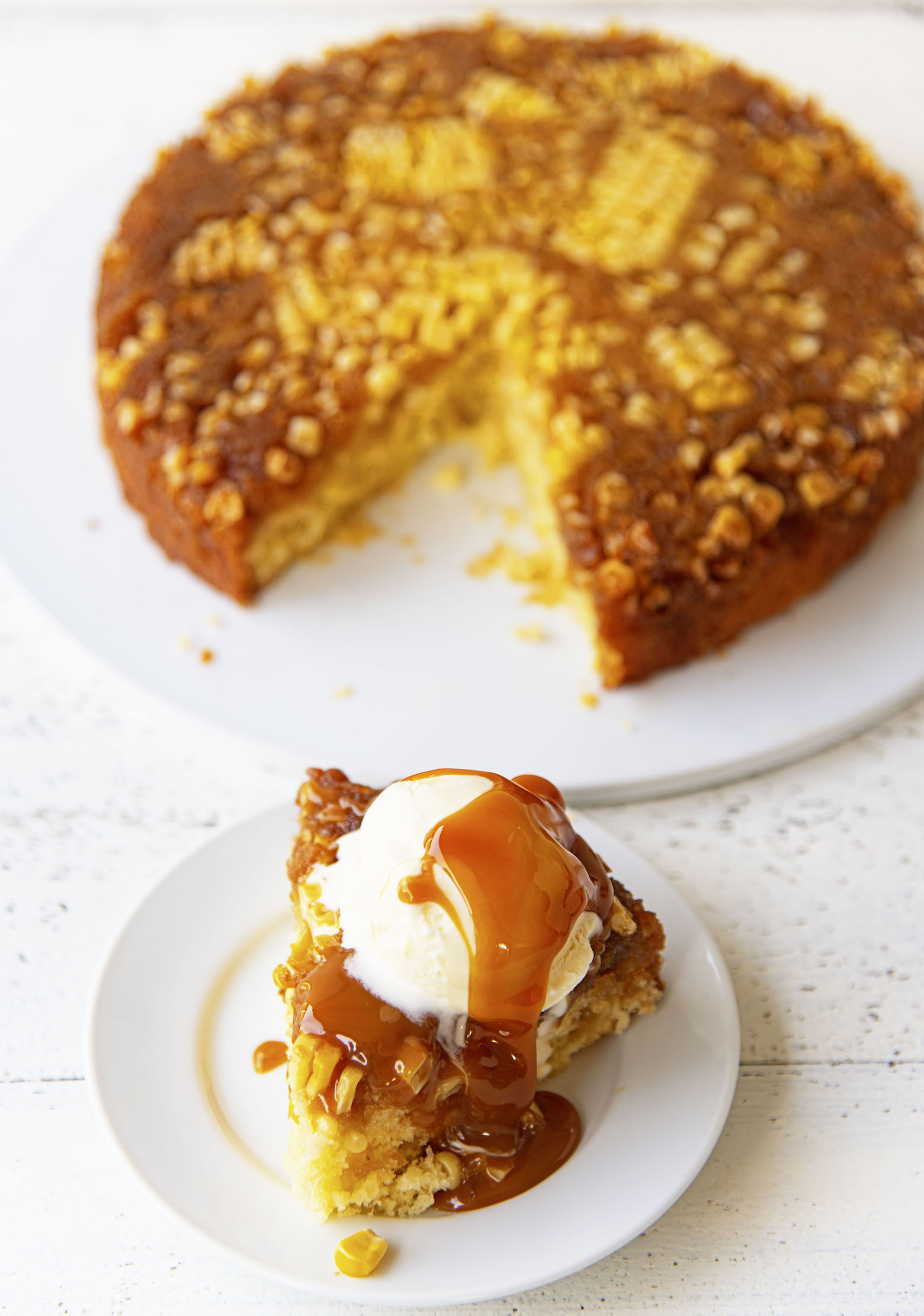 Upside down cake on a small white plate with vanilla ice cream and caramel sauce dripping down. Remainder of cake in the background seen with a slice missing.