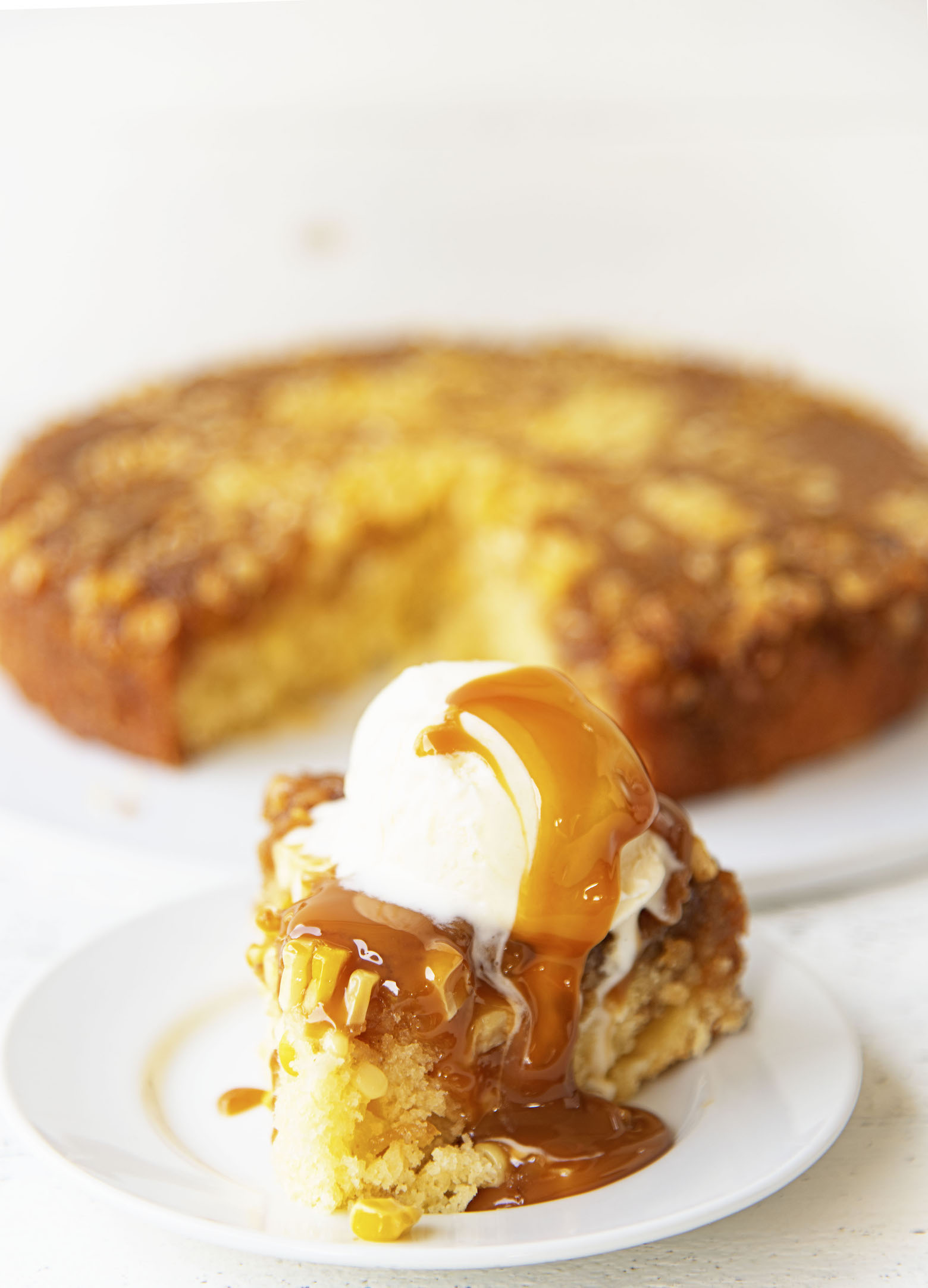 Side view of upside down cake on a small white plate with vanilla ice cream and caramel sauce dripping down. Remainder of cake in the background. 