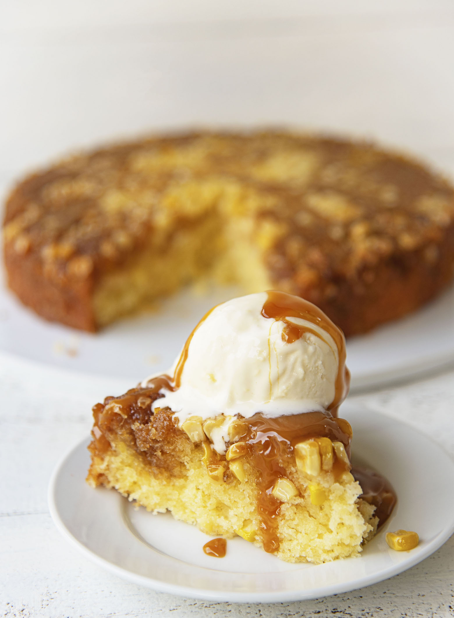 Side view of upside down cake on a small white plate with vanilla ice cream and caramel sauce dripping down. Remainder of cake in the background. 