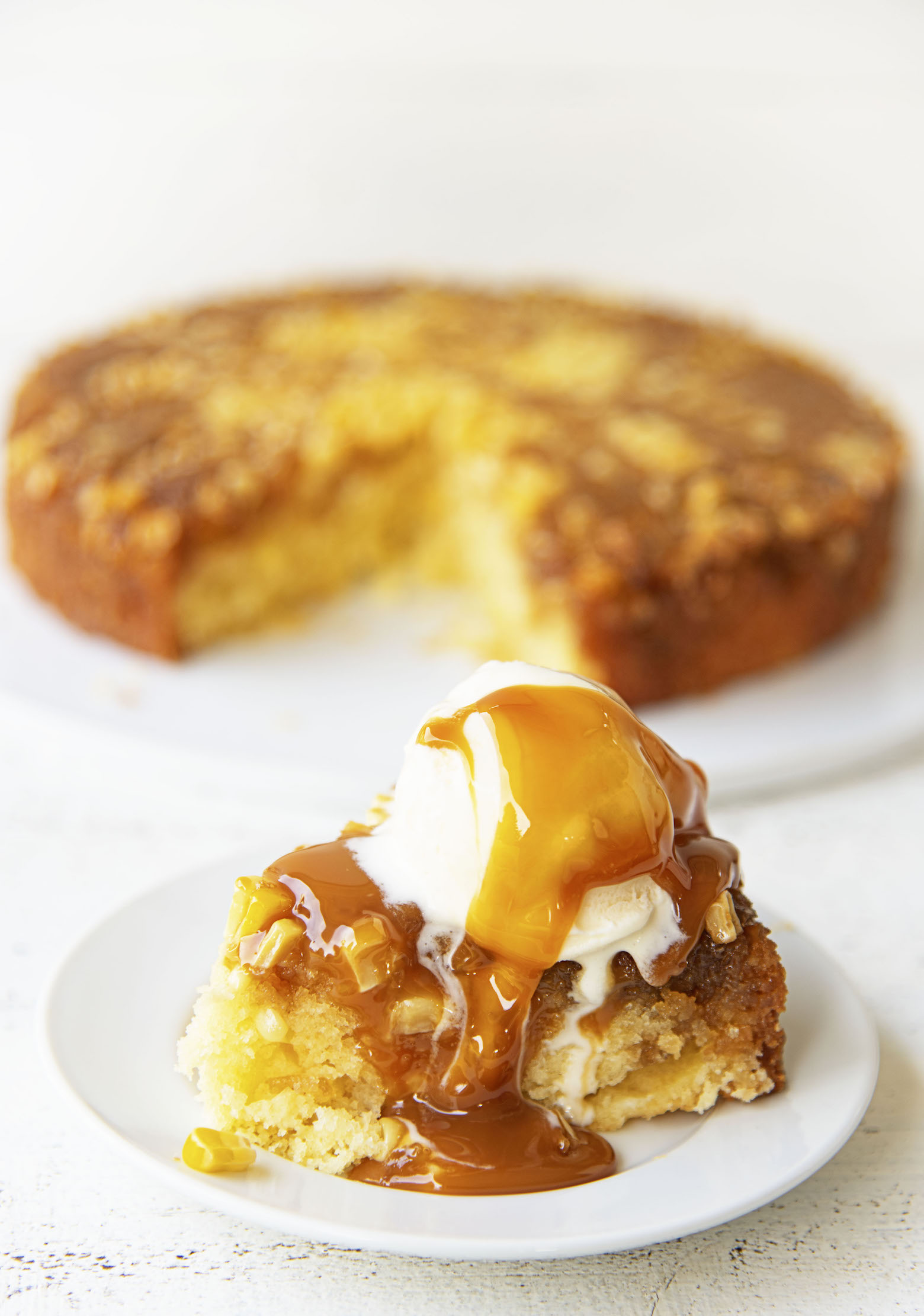Upside down cake on a small white plate with vanilla ice cream and caramel sauce dripping down. Remainder of cake in the background.