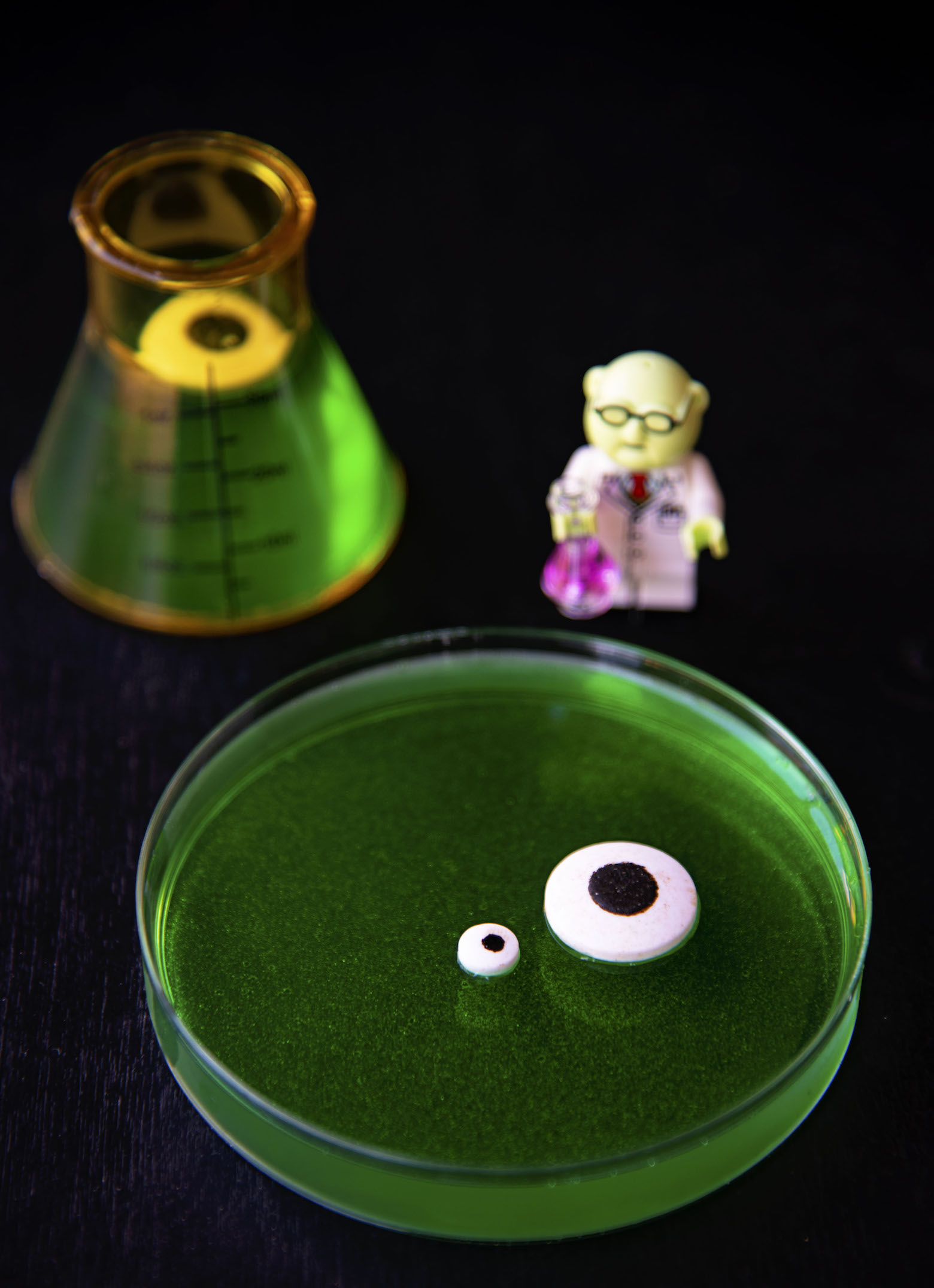 Close up of a single petri dish with green jello and eyeballs next to a beaker with an eyeball and Mr Bunsen.