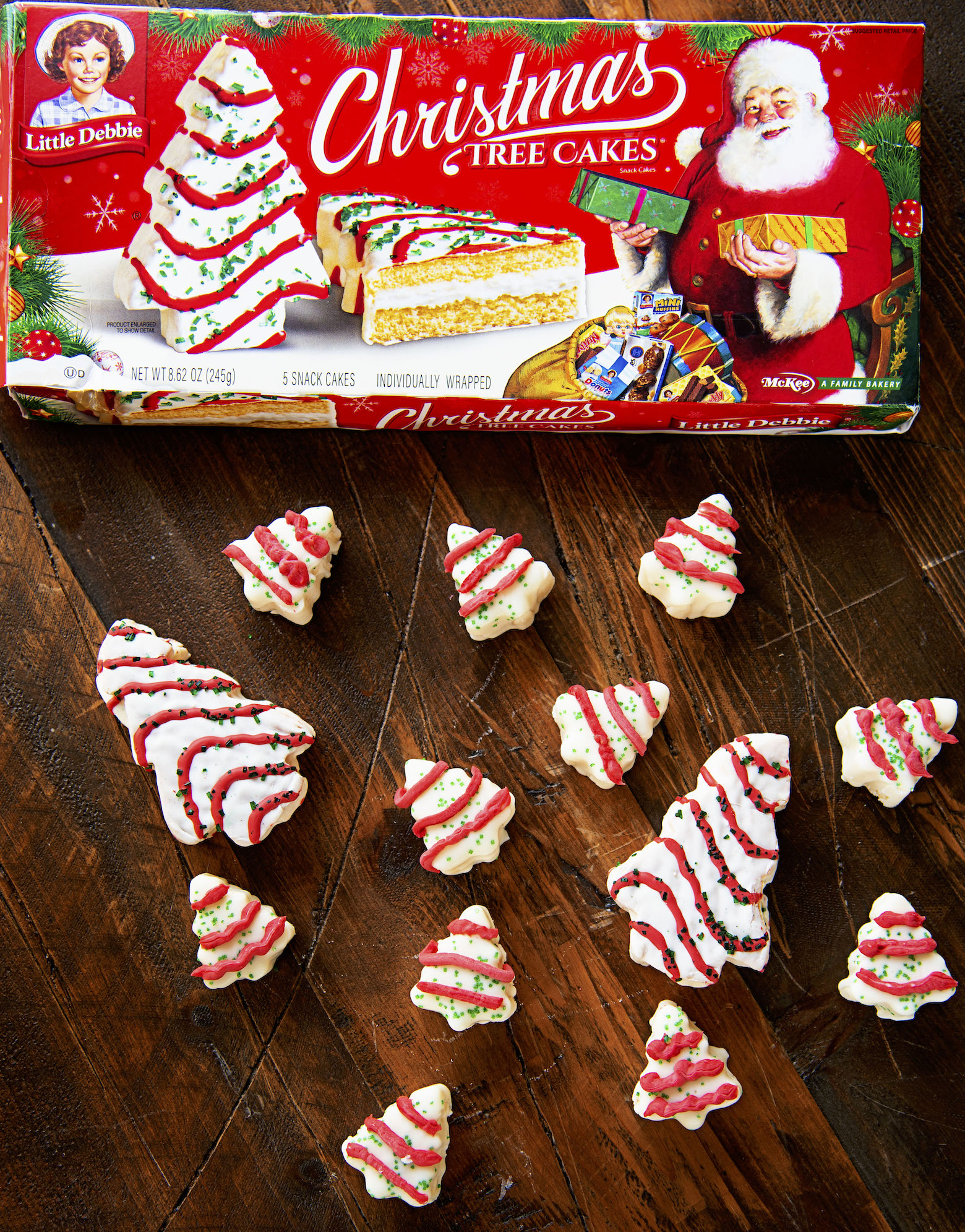 Scattered Christmas Tree Cake Fudge pieces on a wood background with a package of Little Debbie Christmas Tree Cakes box. 