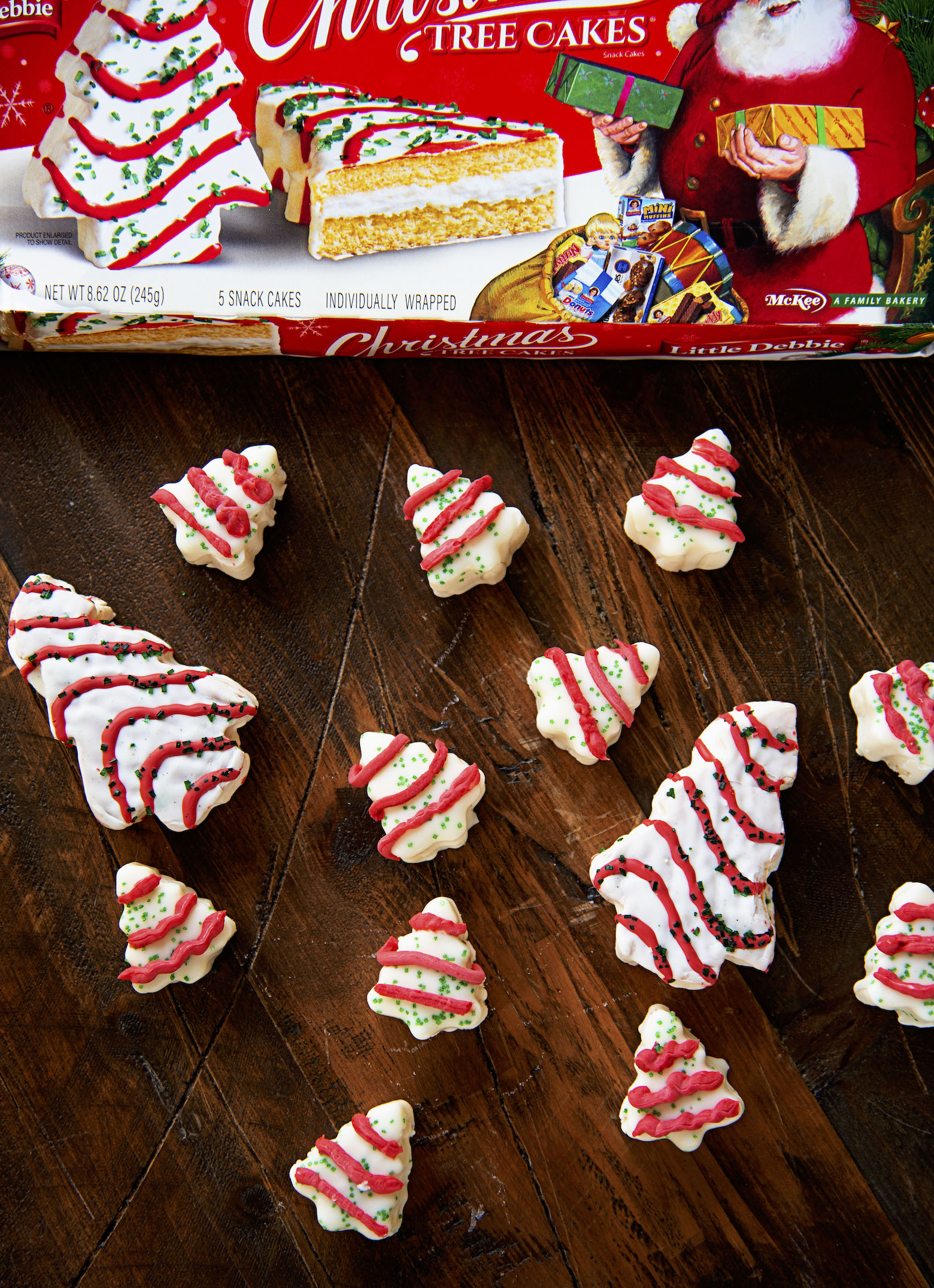 Scattered Christmas Tree Cake Fudge pieces on a wood background with a package of Little Debbie Christmas Tree Cakes box. 