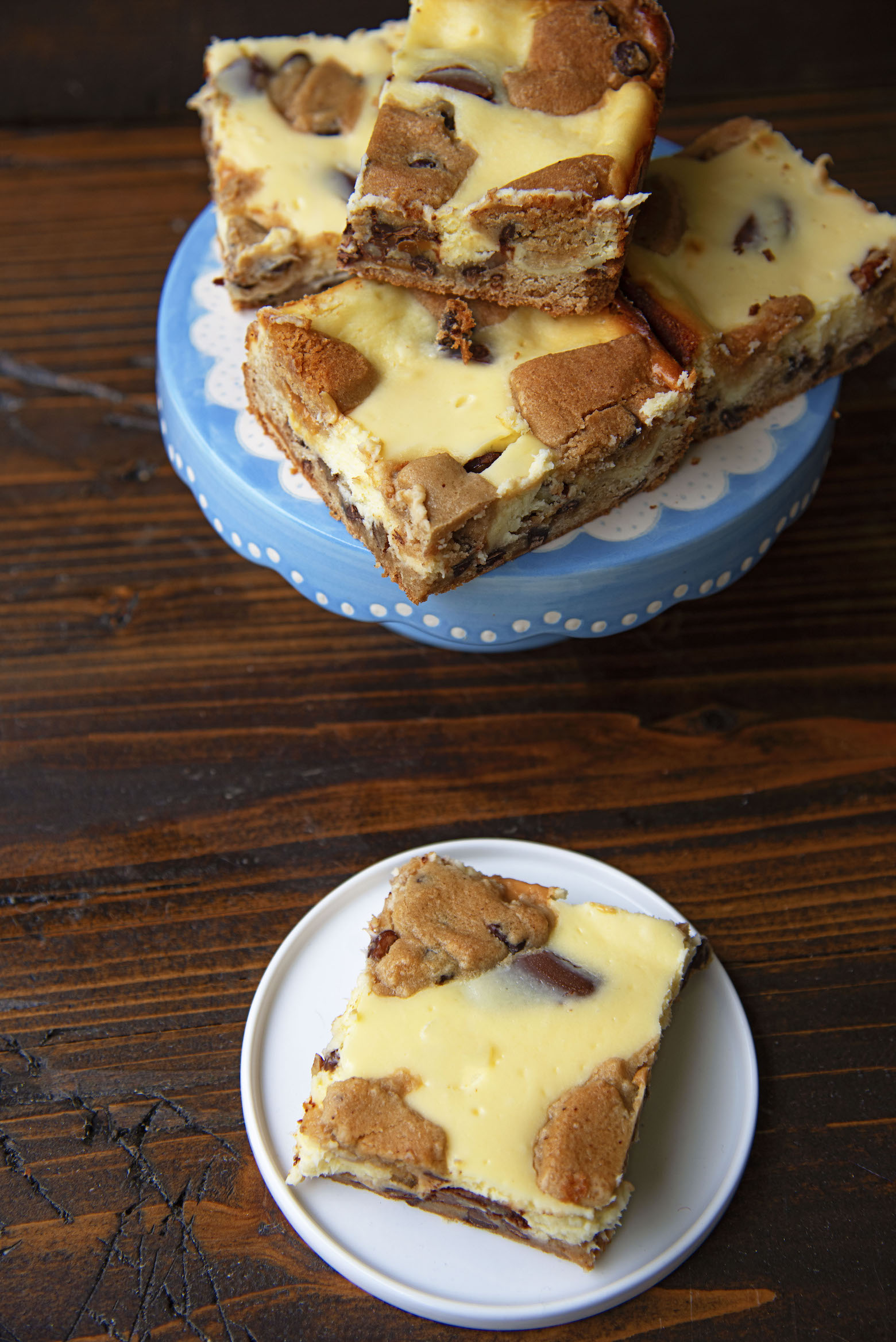 Caramel Turtle Cheesecake Bar on a small white plate with remaining bars on a cake stand in the background. 