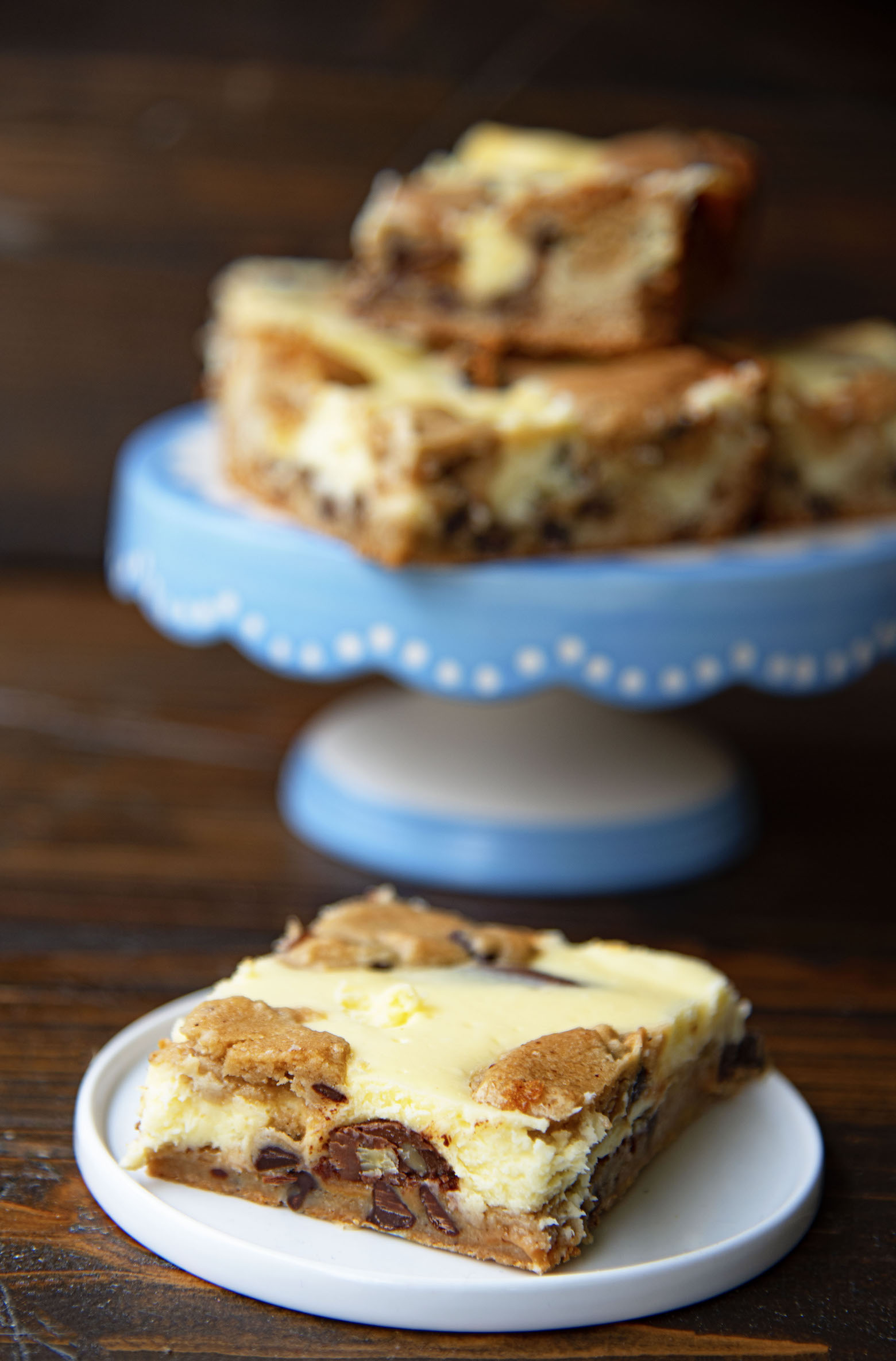 Caramel Turtle Cheesecake Bar on a small white plate with remaining bars on a cake stand in the background. 