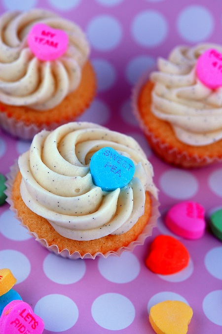 Three Creamsicle Conversation Heart Cupcakes on pink polka dot background with candy hearts sprinkled around them. 