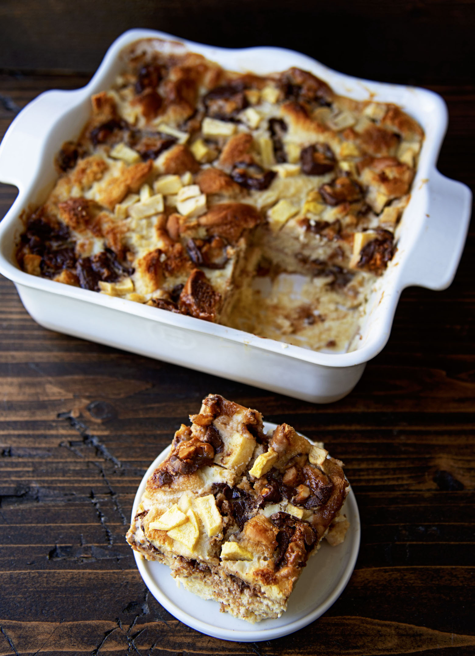 Single bread pudding slice on a small plate in front with remaining amount of bread pudding in a dish behind it. 