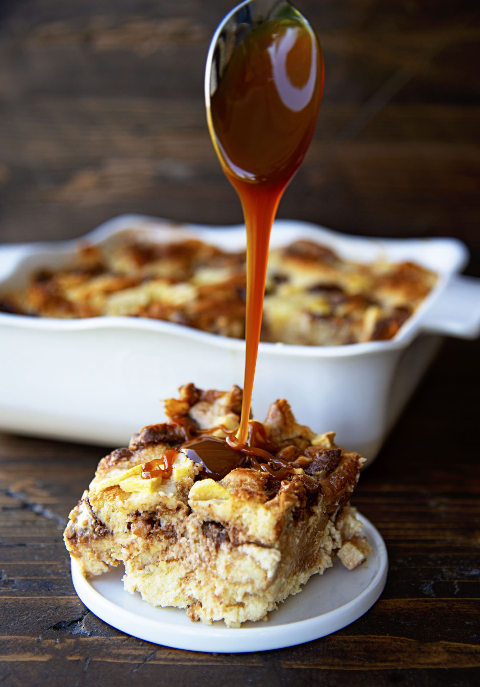 Caramel sauce falling from a spoon onto a slice of bread pudding. 