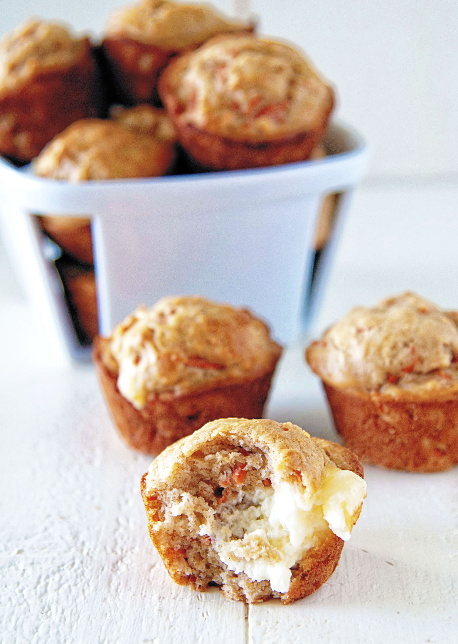 Three muffins in front of a small basket of muffins. The first muffin has a bite out of it exposing the cream cheese frosting middle.
