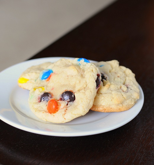 Small white plate on a brown surface with three cookies on the plate with colorful m and m's. 