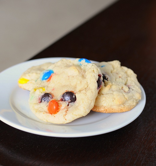 Small white plate on a brown surface with three cookies on the plate with colorful m and m's.
