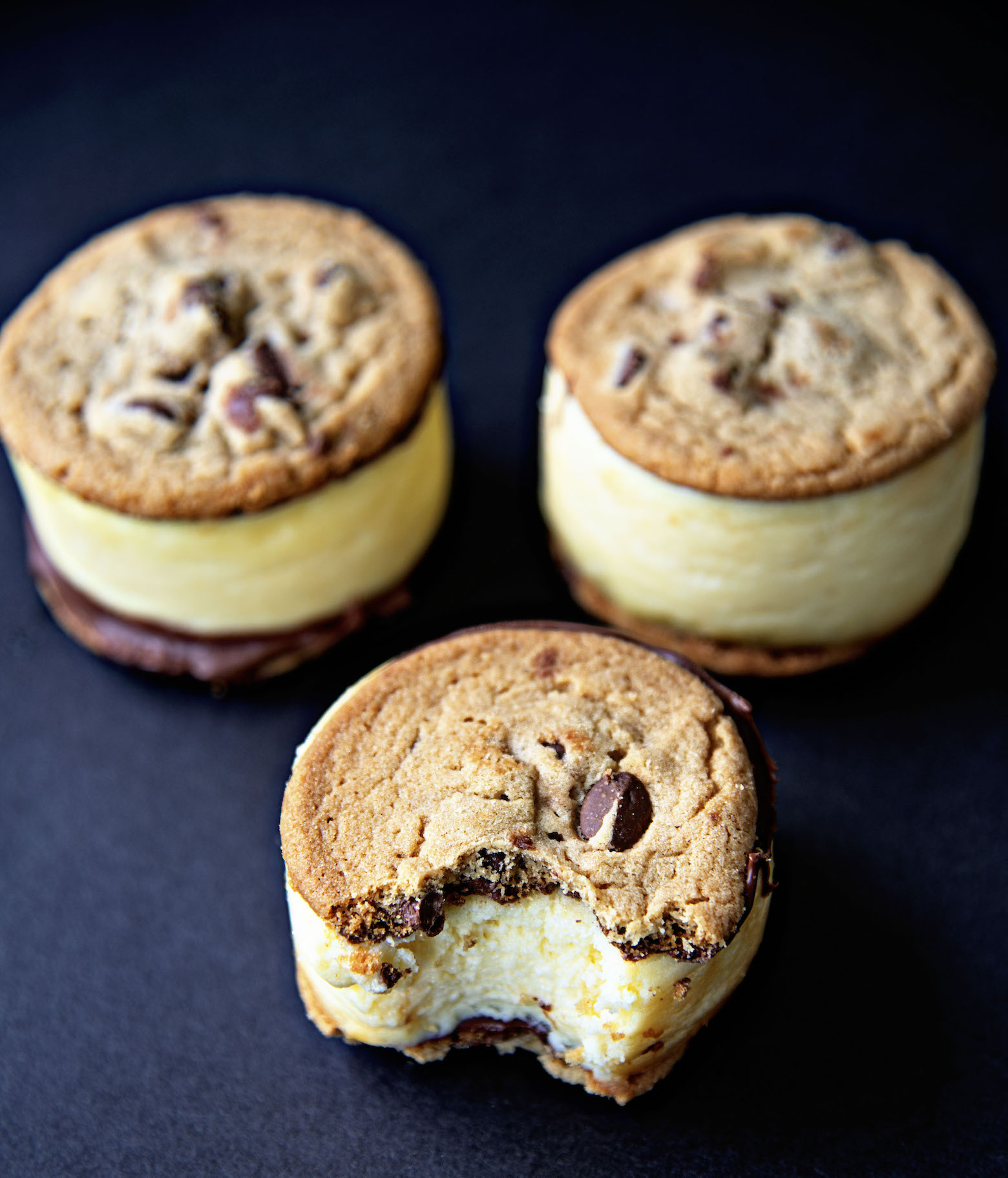 Three Chocolate Chip Cookie Cheesecake Sandwiches staggered with the front one having a bite out of it. Black background. 