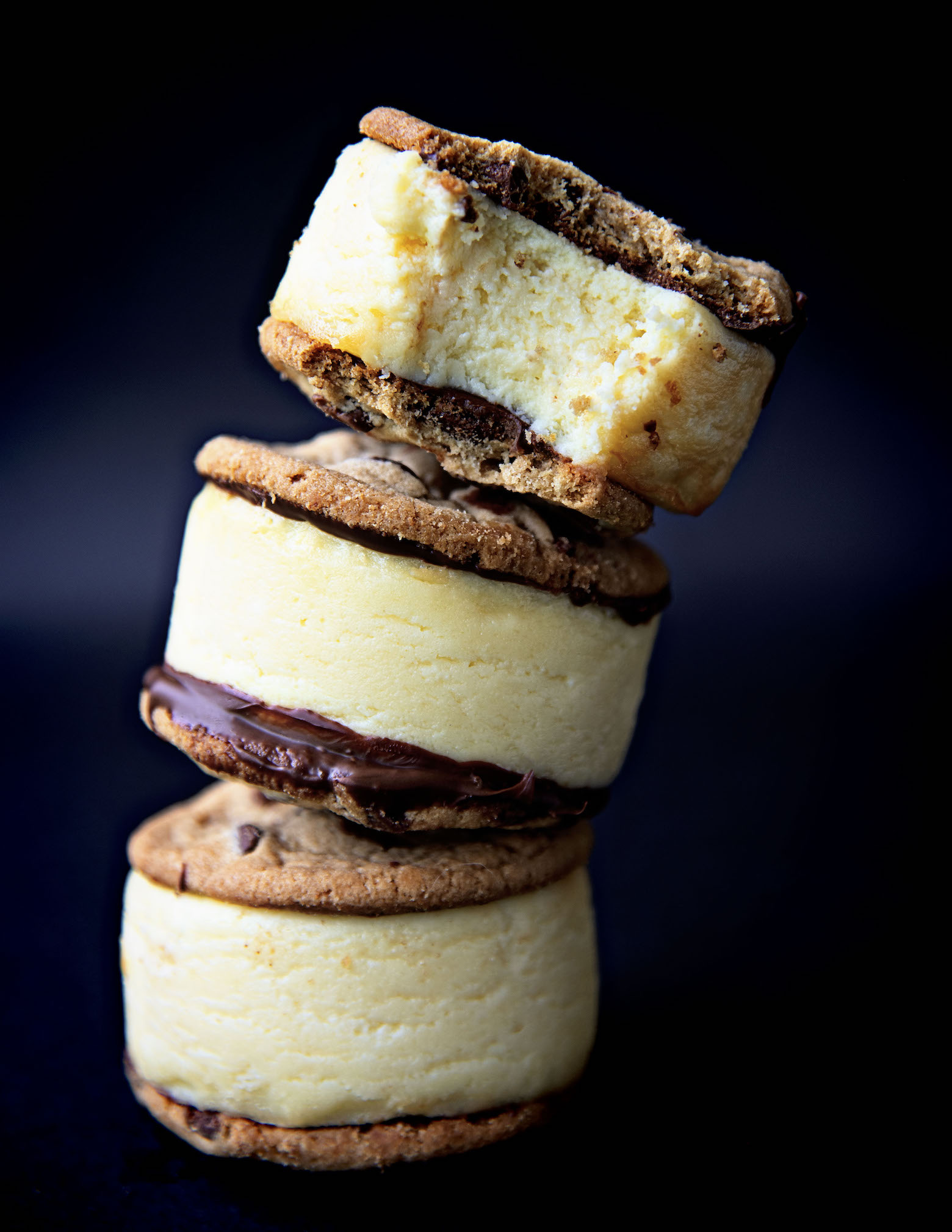 Stack of three Chocolate Chip Cookie Cheesecake Sandwiches with the top one having a bite out of it. Background is black.