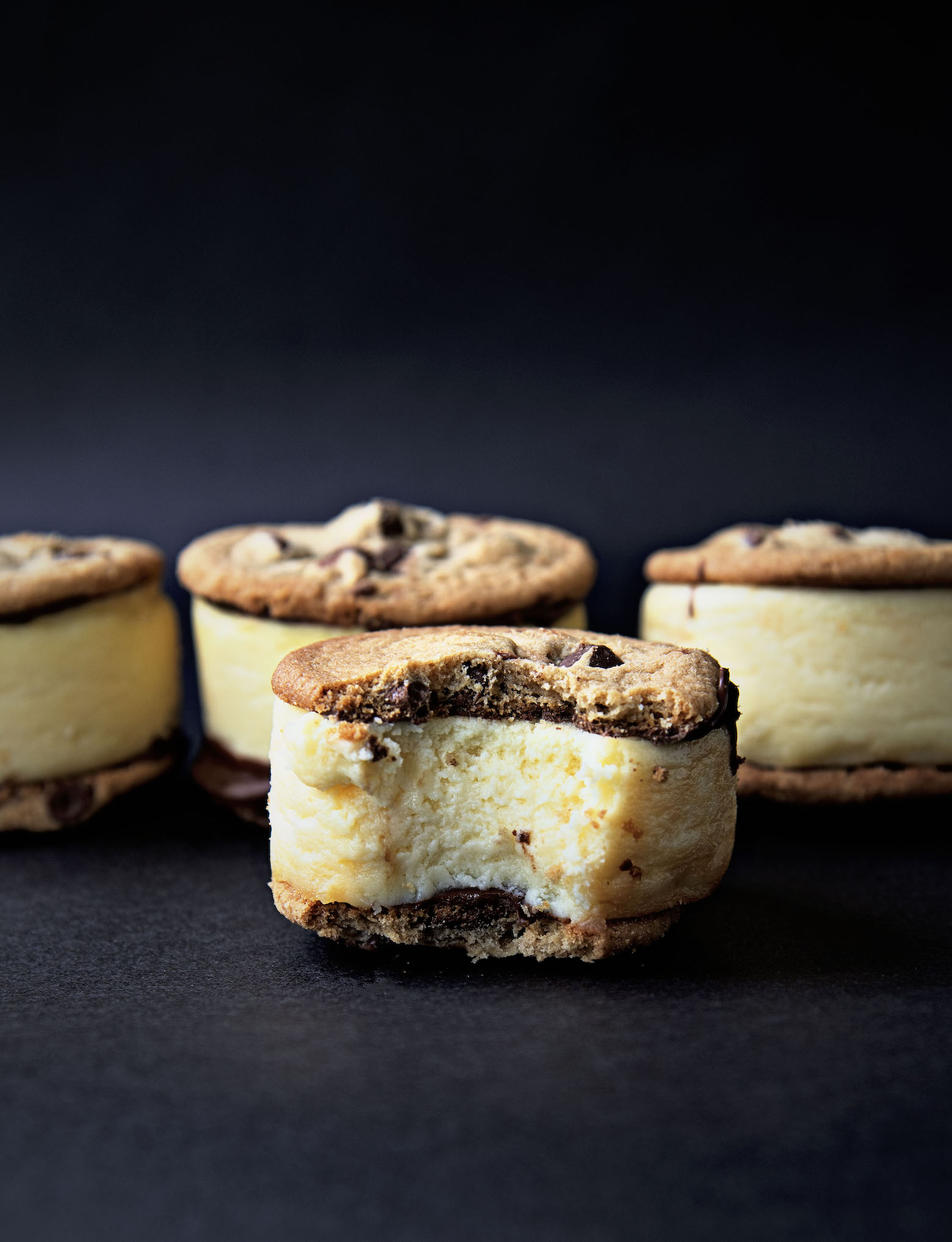 Several Chocolate Chip Cookie Cheesecake Sandwich in a row with one sandwich with a bite out of it in front of the others. Black background.