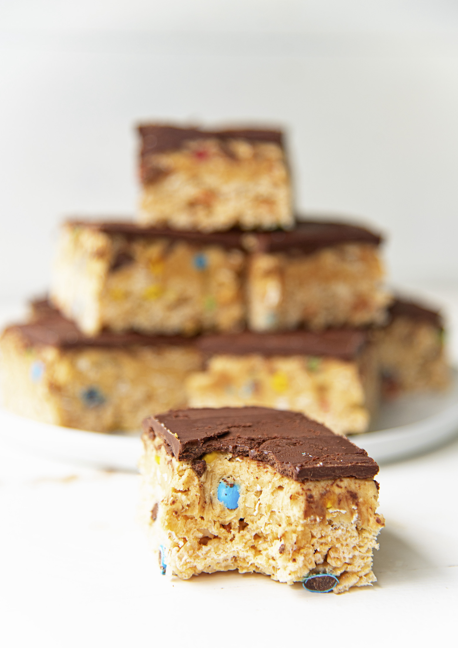 A single Monster Cookie Dough Rice Krispie Treat with a bite out of it in front of a plate stacked with more treats. 