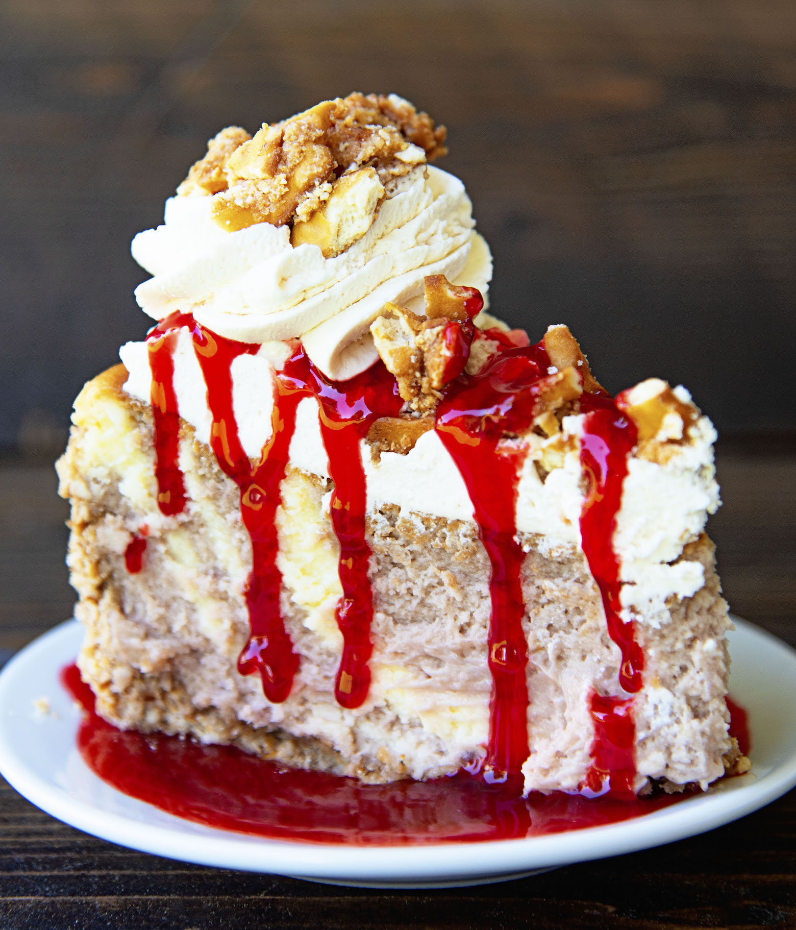 Slice of Strawberry Pretzel Crusted Cheesecake on a small white plate in a pool of strawberry sauce with strawberry sauce dripping down the side of the cheesecake. 