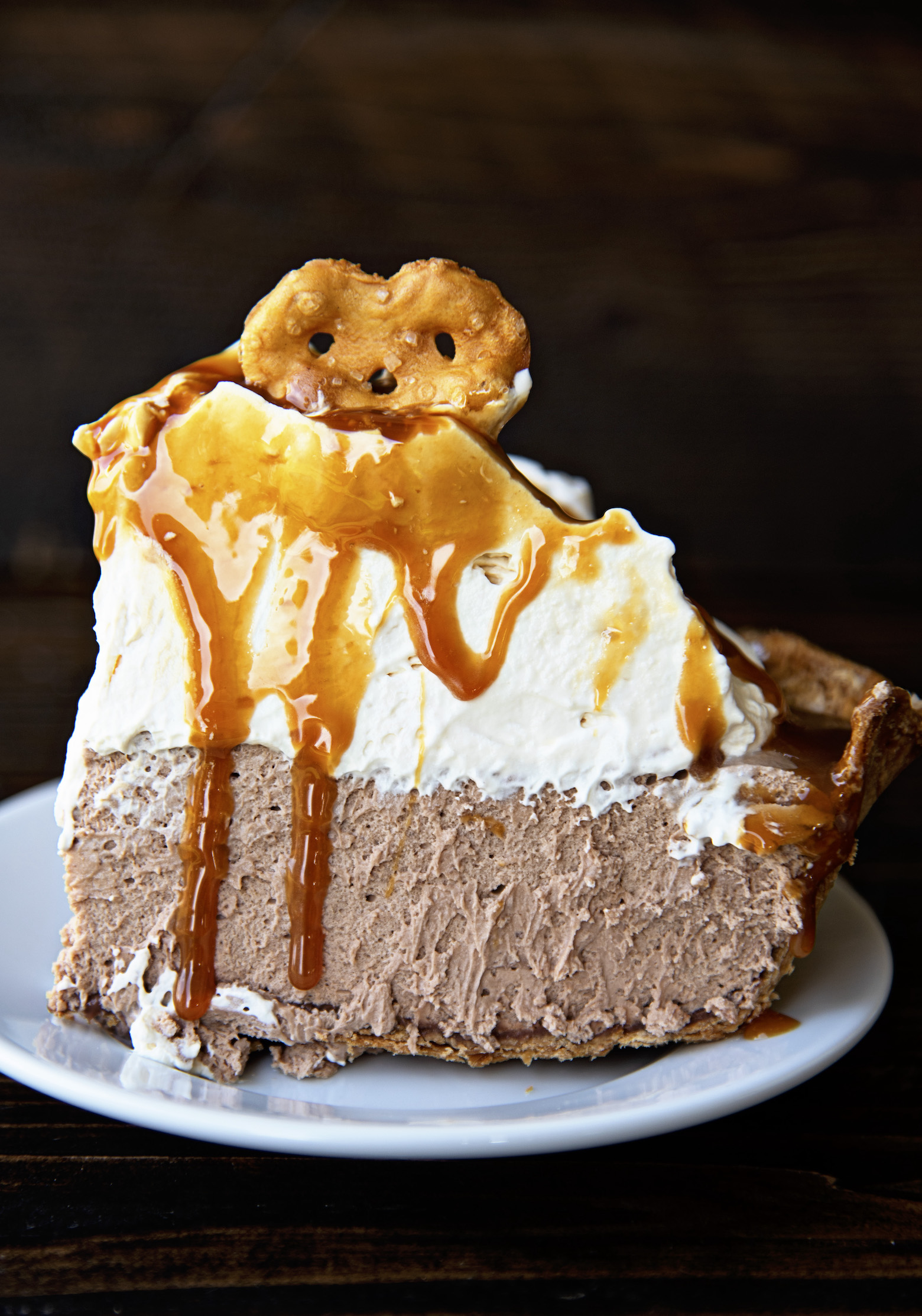 Close up of a single slice of pie from a side view. You can see the pretzel crust, chocolate caramel filling, tall whipped cream, caramel sauce dripping down, and the mini pretzel on top. 