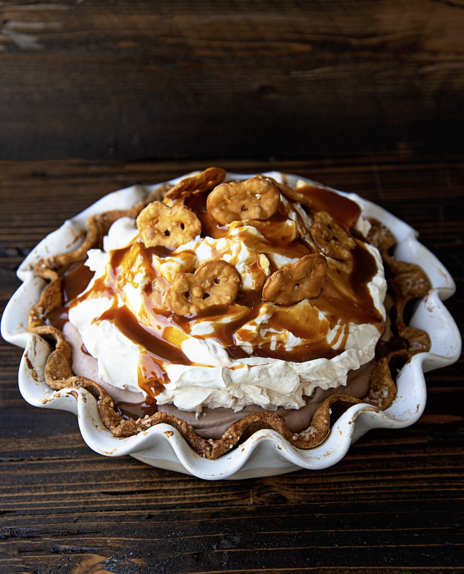 The full pie still in it's white pie pan. You can see the chocolate filling peaking through with whipped cream, caramel and pretzels on top.