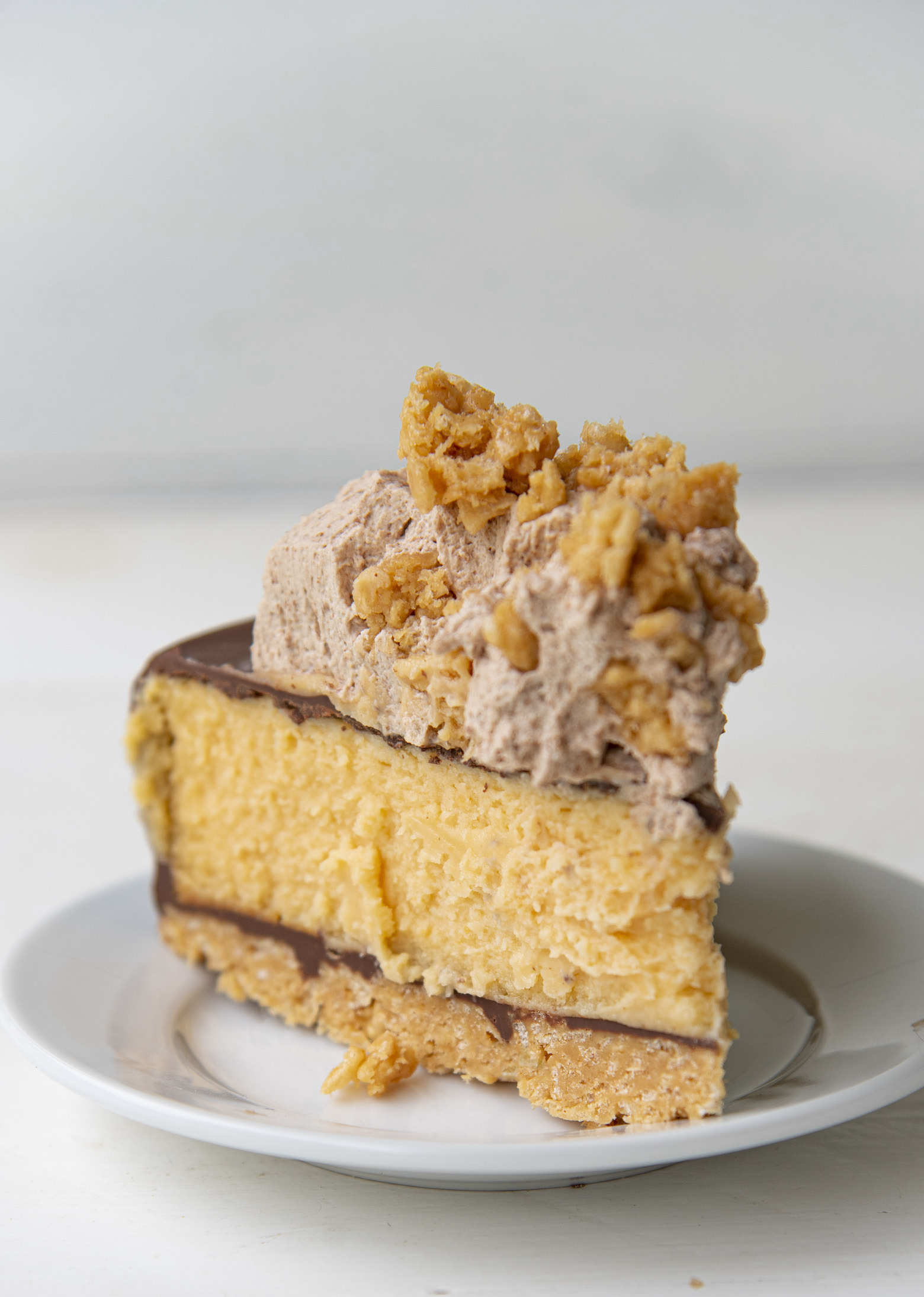 Slice of the Chocolate Glazed Scotcharoo Cheesecake on a small white plate. With focus on the chocolate butterscotch whipped cream and cereal pieces on top. 