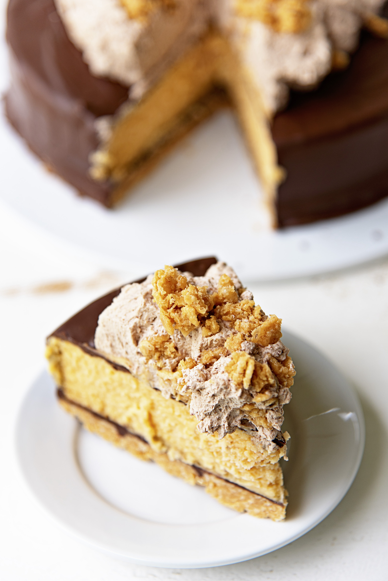 Three quarter view of the a single slice of cheesecake with the focus on the top of the cheesecake which is chocolate butterscotch whipped cream and cereal pieces. 