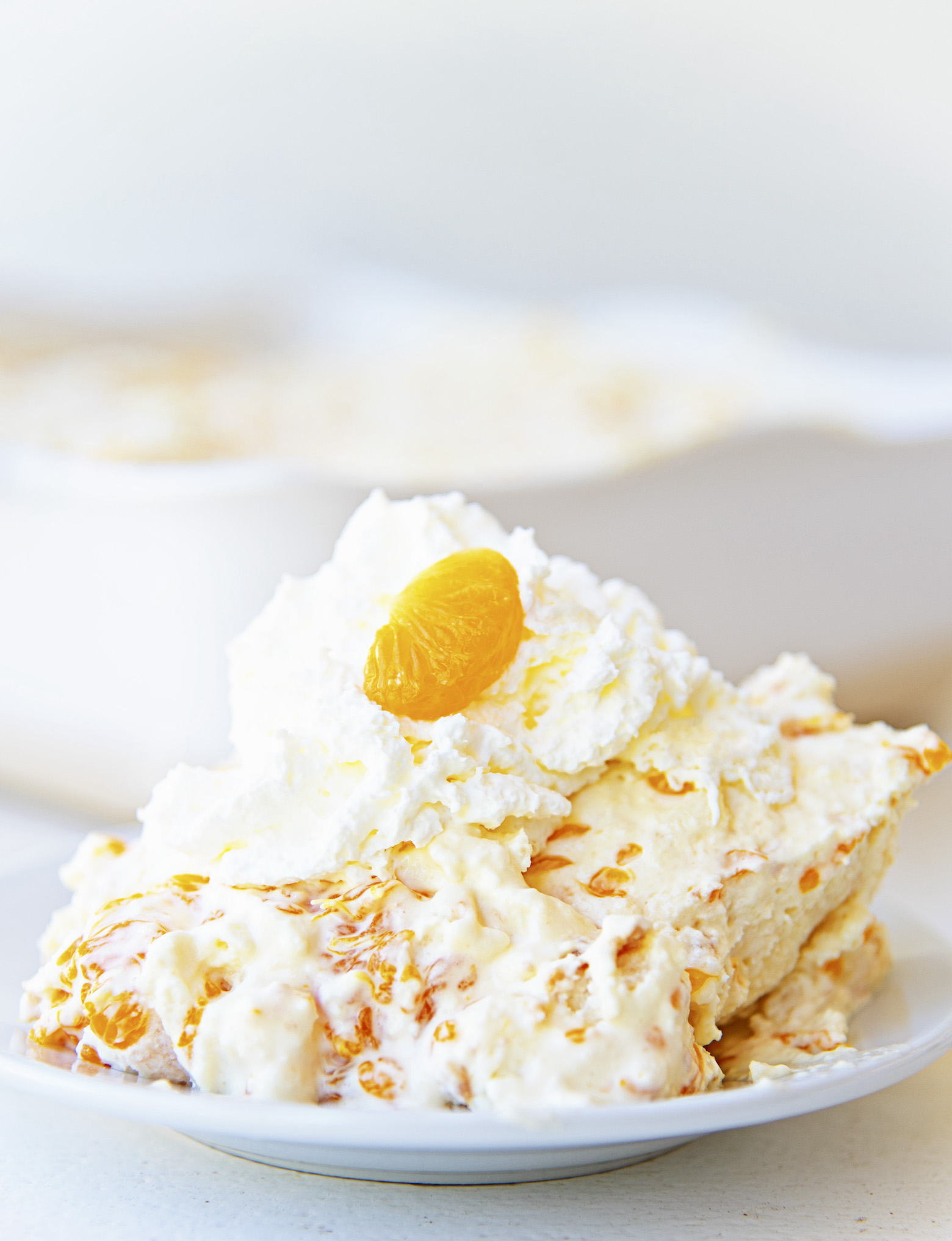 Scoop of angel dessert on a small white plate with whipped cream and a small orange slice on top with remaining dessert in back. 