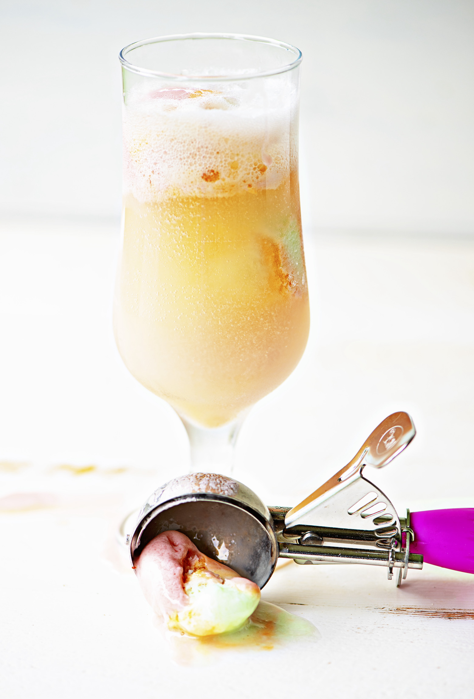 Single glass with bubbles from the selzer in the cocktail with scoops of rainbow sherbet in it. A ice cream scoop with some rainbow sherbet still in it sits below the glass. 