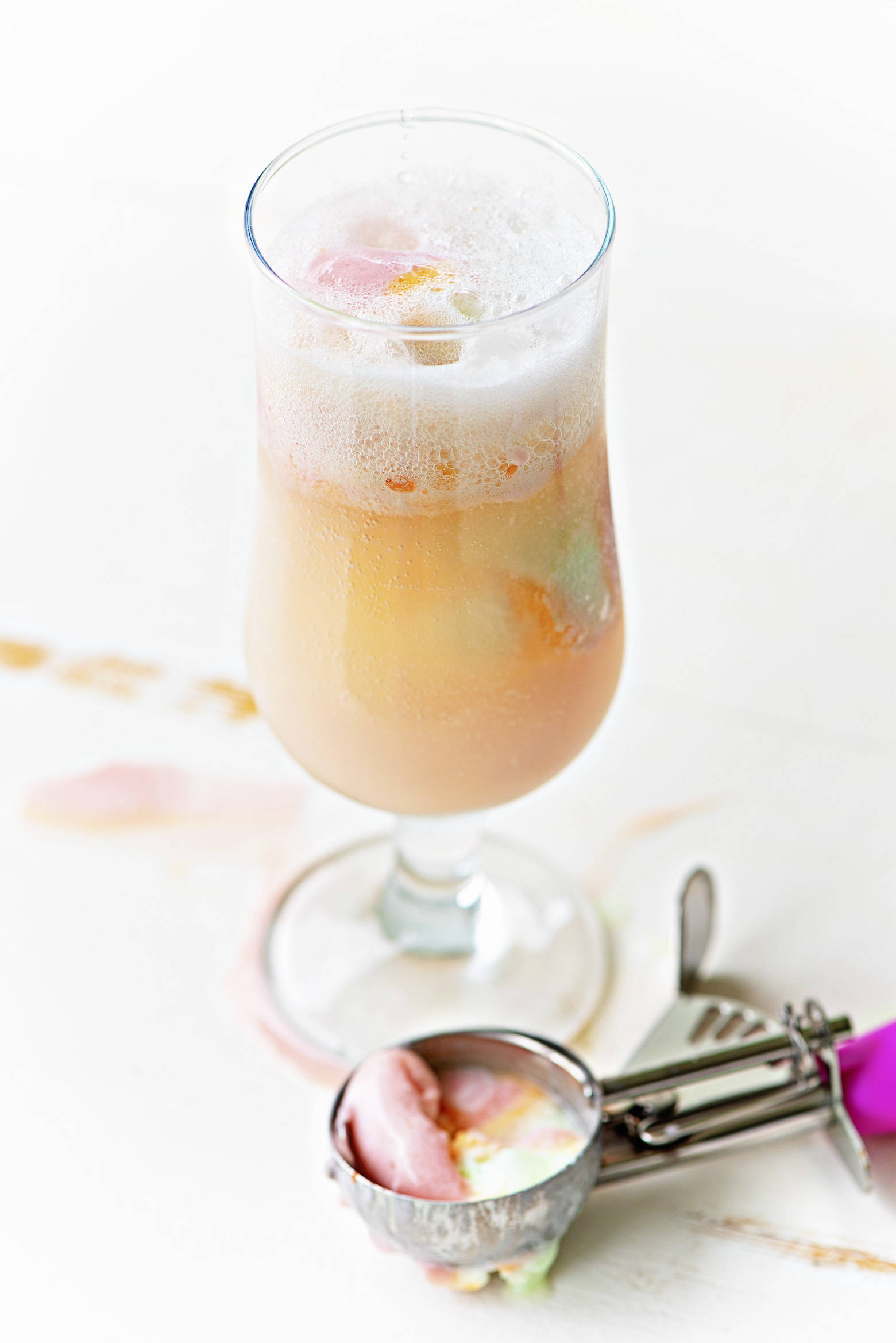 Single glass with bubbles from the selzer in the cocktail with scoops of rainbow sherbet in it. A ice cream scoop with some rainbow sherbet still in it sits below the glass.