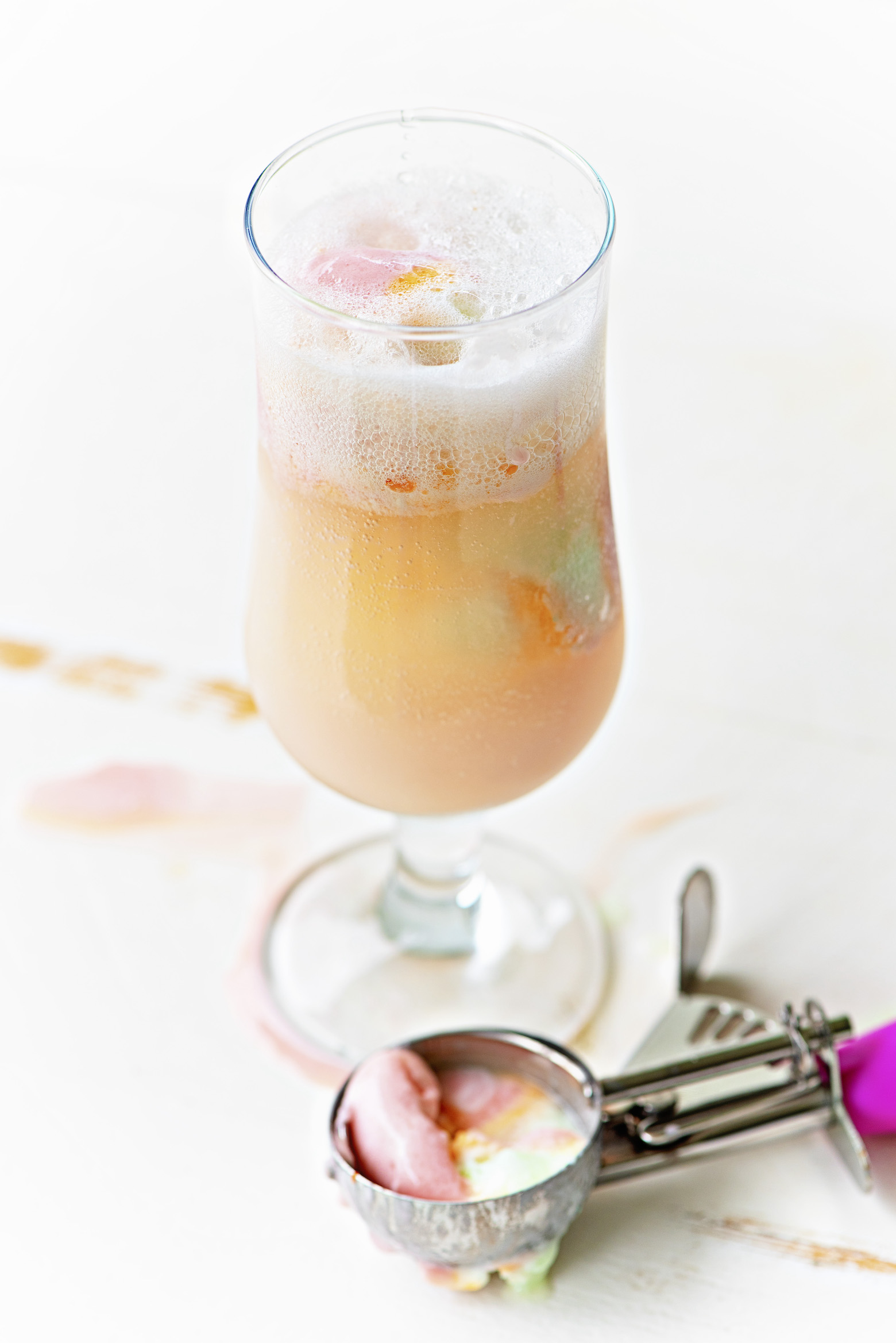 Single glass with bubbles from the selzer in the cocktail with scoops of rainbow sherbet in it. A ice cream scoop with some rainbow sherbet still in it sits below the glass. 