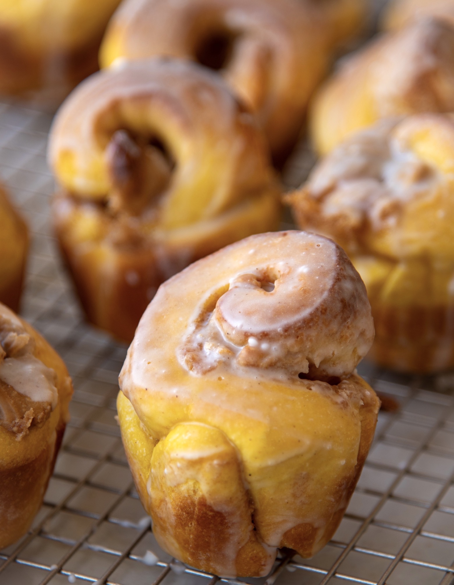 Several Pumpkin Cinnamon Crunch Buns on a wire rack with glaze dripped below. Focus is on one bun. 