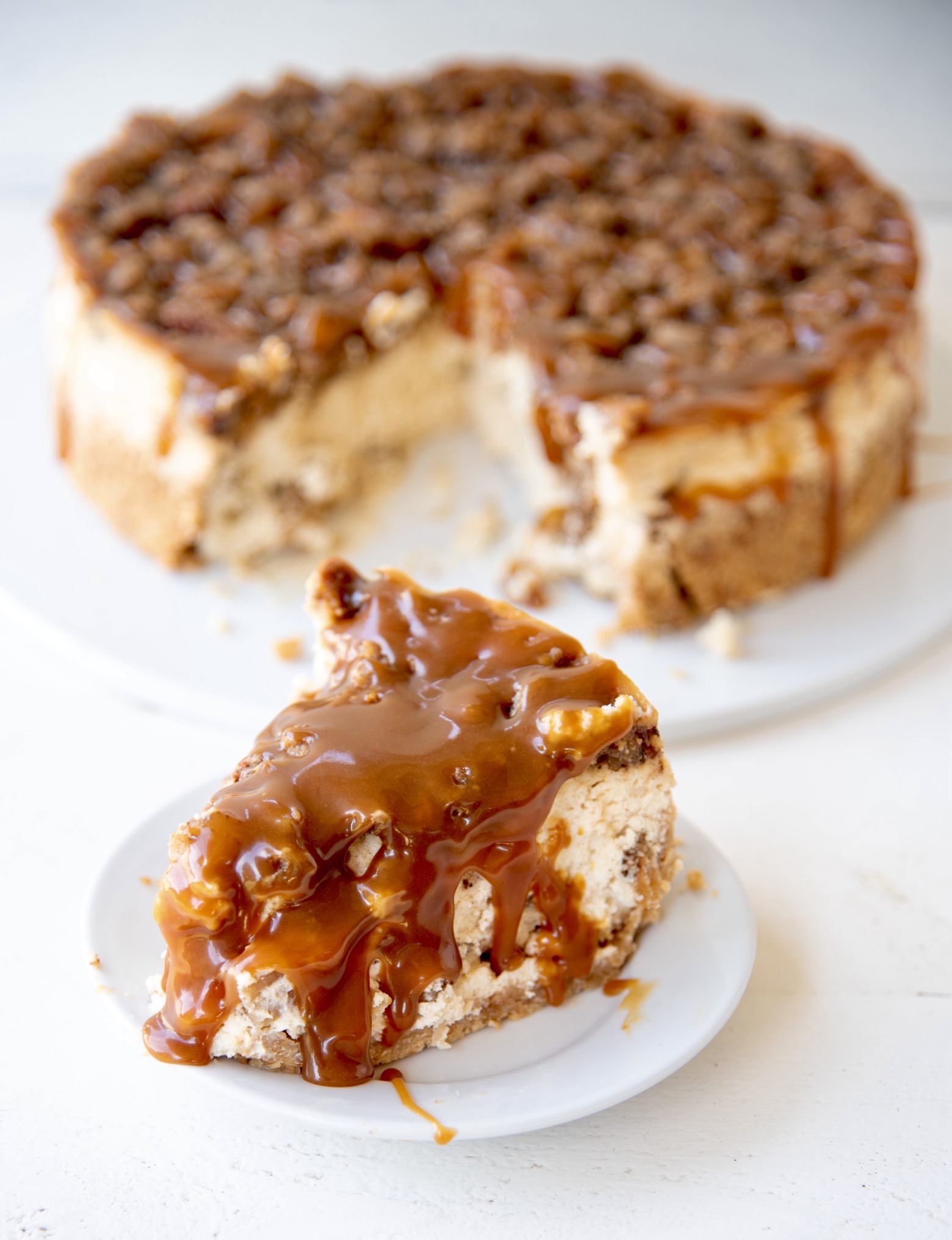 Piece of cheesecake on a small white plate with the remaining cheesecake in back with caramel sauce drizzled over it.
