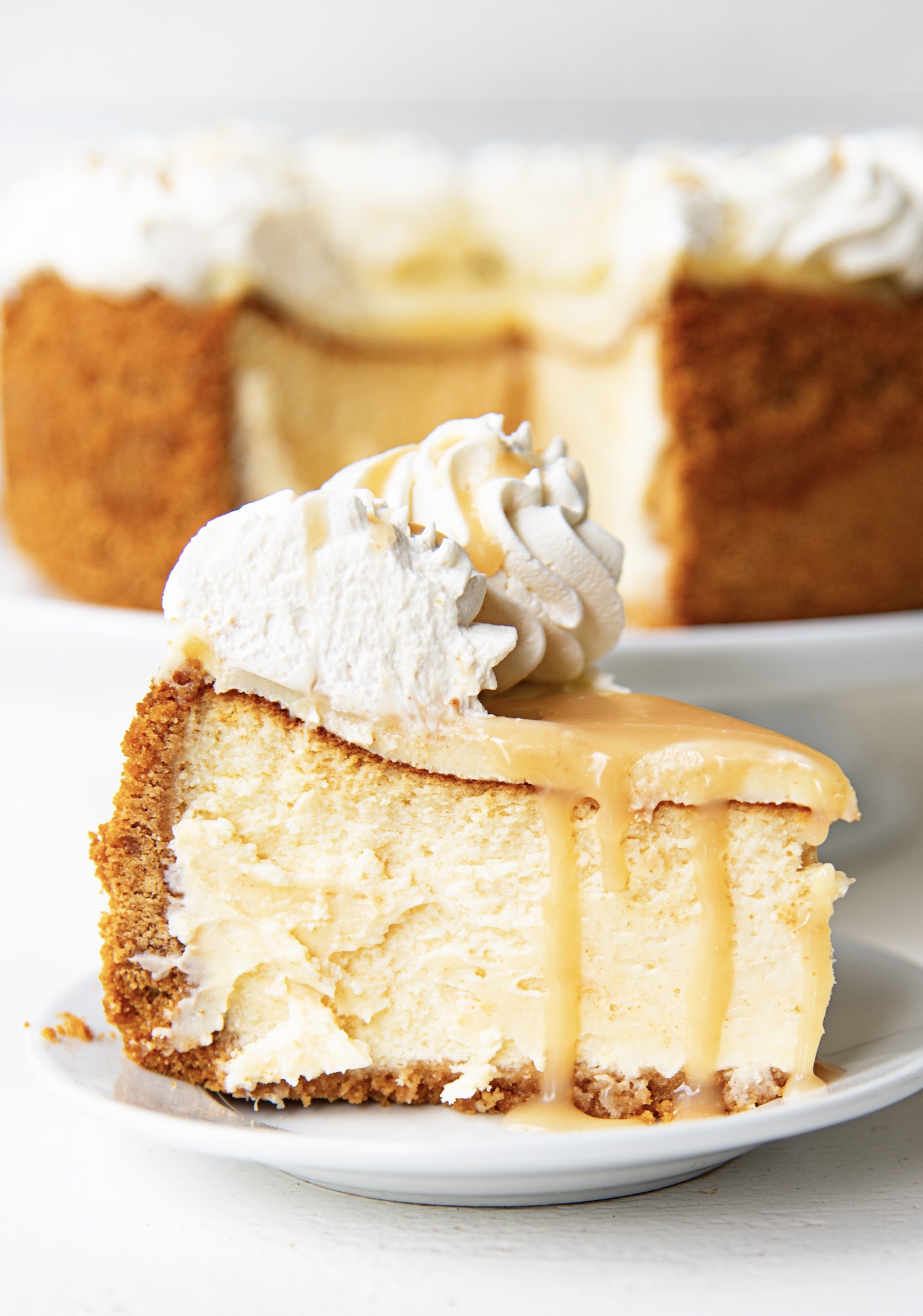 Slice of cheesecake on a small white plate with caramel dripping down the sides with remaining cheesecake in the background. 