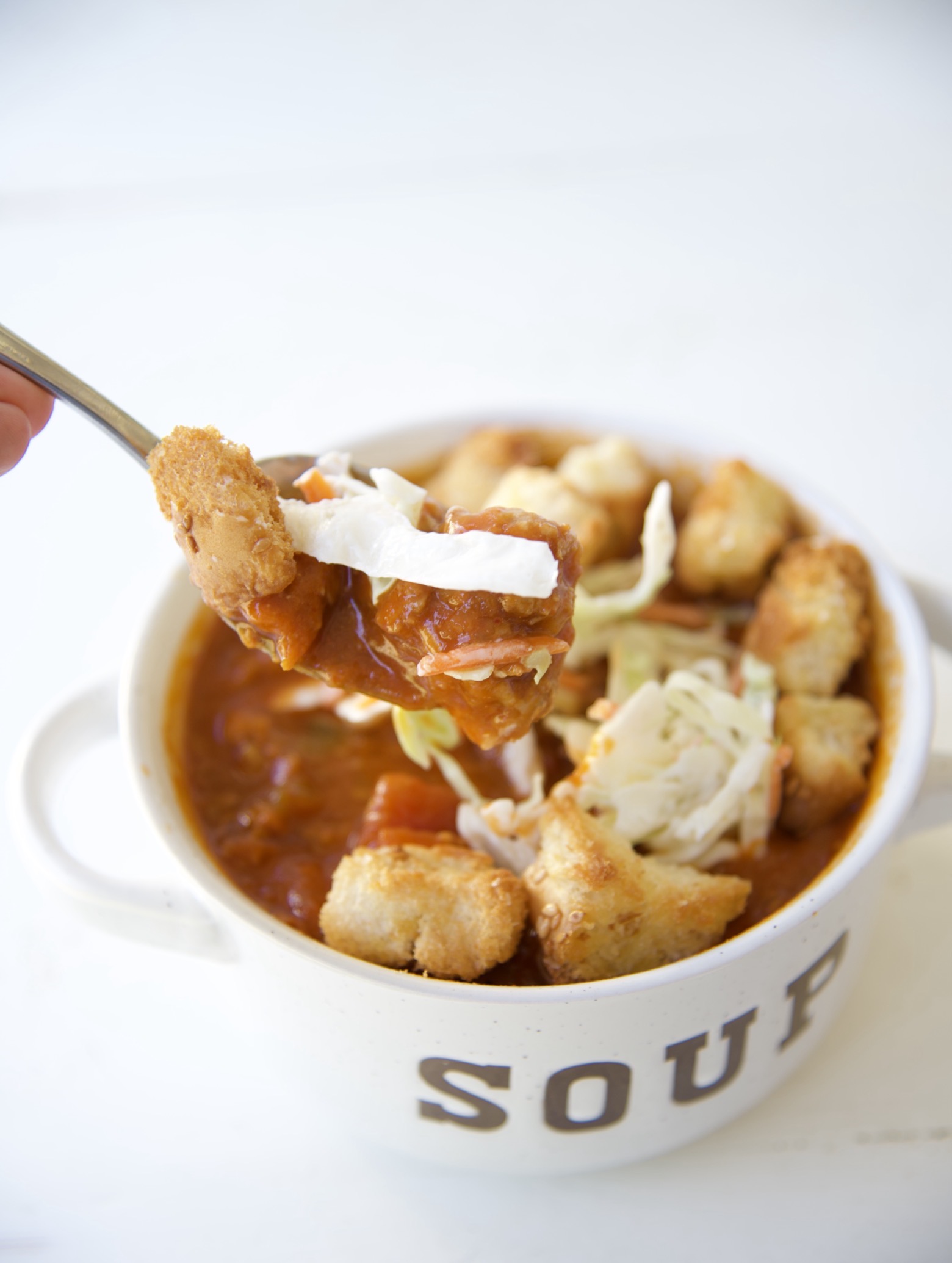 Close up of a spoon with Sloppy Joe Soup on it with coleslaw and croutons in the spoon as well. A full soup bowl is in the background. 