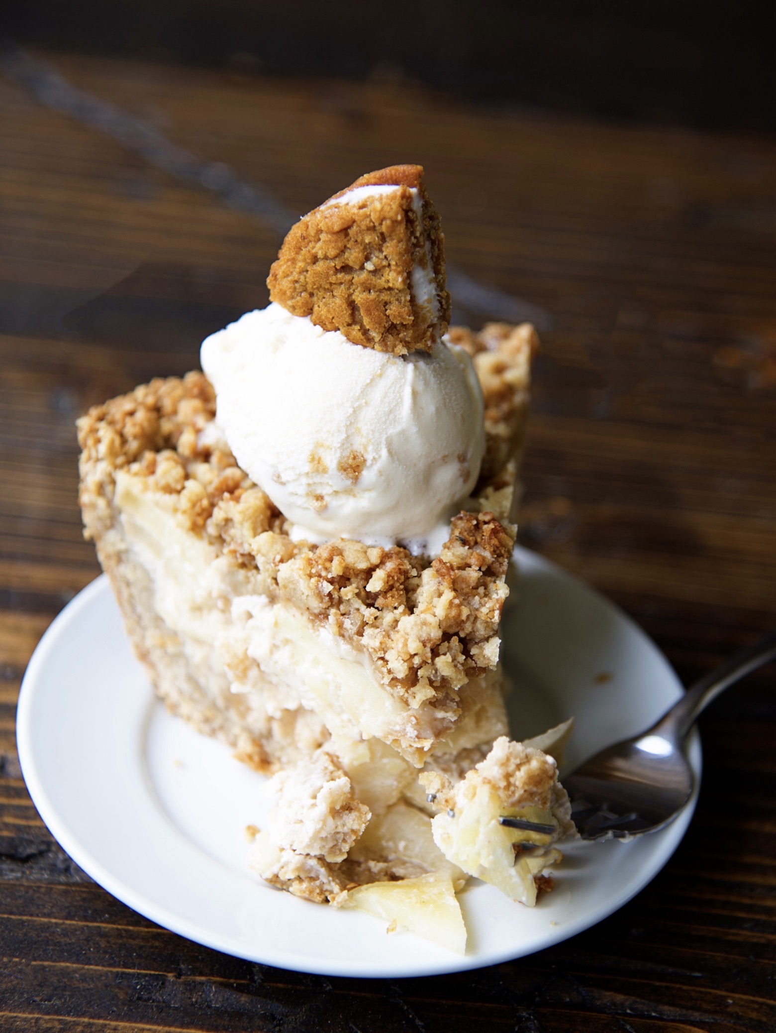 A piece of the Oatmeal Creme Apple Pie on a small white plate with ice cream and a cookie pie wedge on top of the pie with a fork having taken a piece of the pie lays on the plate. 