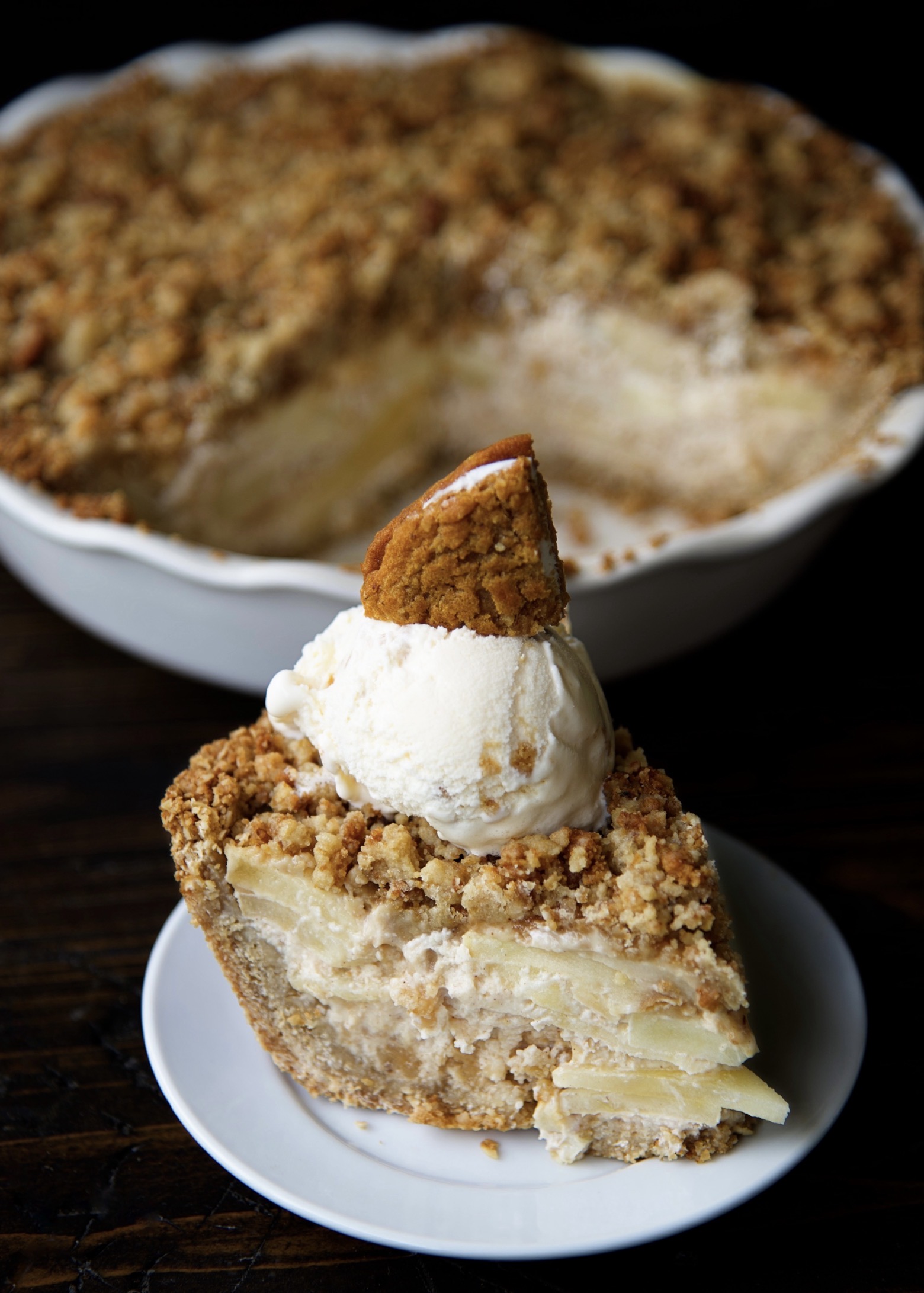 A piece of the Oatmeal Creme Apple Pie on a small white plate with ice cream and a cookie pie wedge on top of the pie. The remaining pie is in the background. 