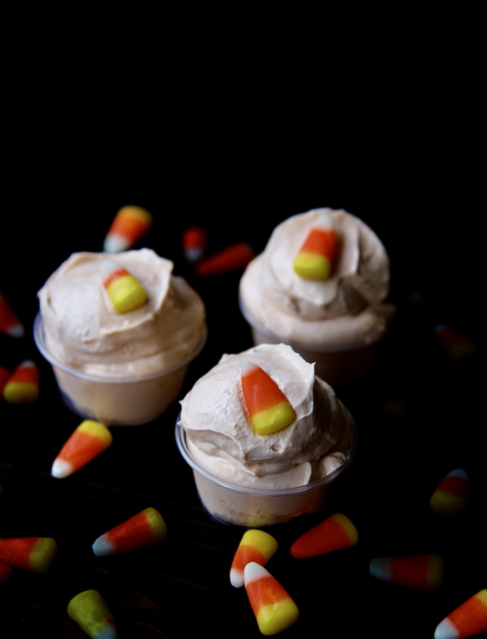 Three quarter view of three pudding shots with a piece of candy corn on each one. With candy corn scattered around.